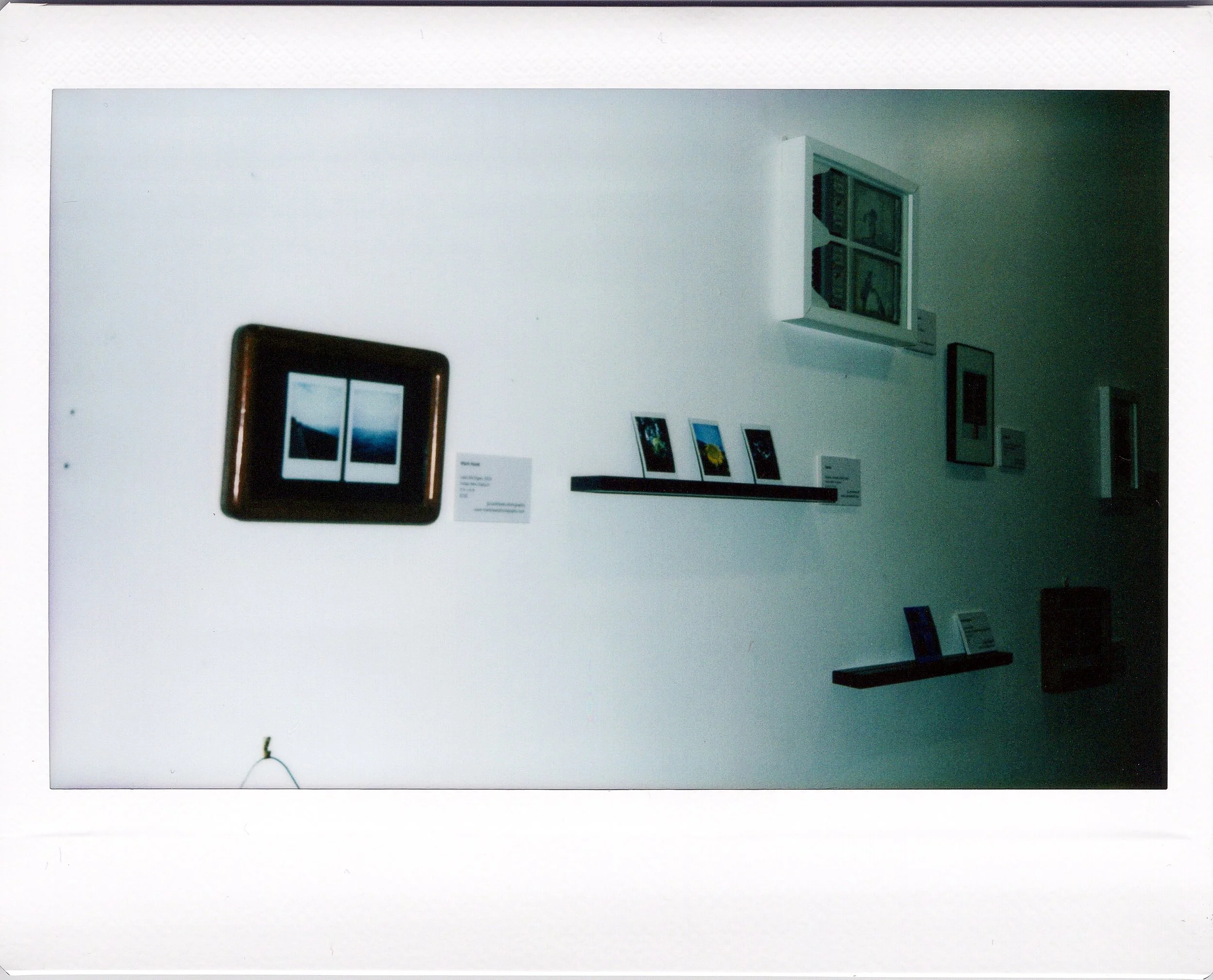 Instant photo of gallery wall with framed polaroid photographs, small photos on a shelf and some artwork, displayed in an art exhibition.