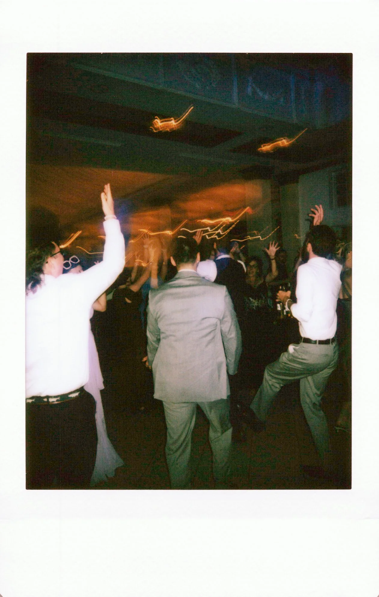 People dancing at a party or nightclub, with some raising their hands, in a dimly lit room with colorful lights and a bar in the background.