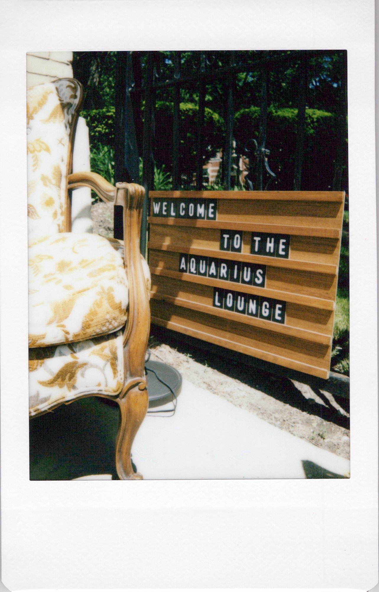 A vintage upholstered armchair with a floral pattern on a white background. Behind it is a wooden sign with black and white letters that says "Welcome to the Aquarius Lounge." In the background, there is a black metal fence, greenery, and trees.