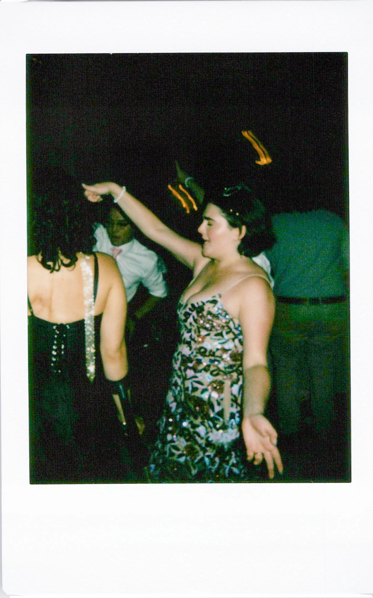 A group of people dancing at a party or nightclub, with a woman in the foreground wearing a patterned dress and raising her arm, and others blurred in the background under dark lighting.