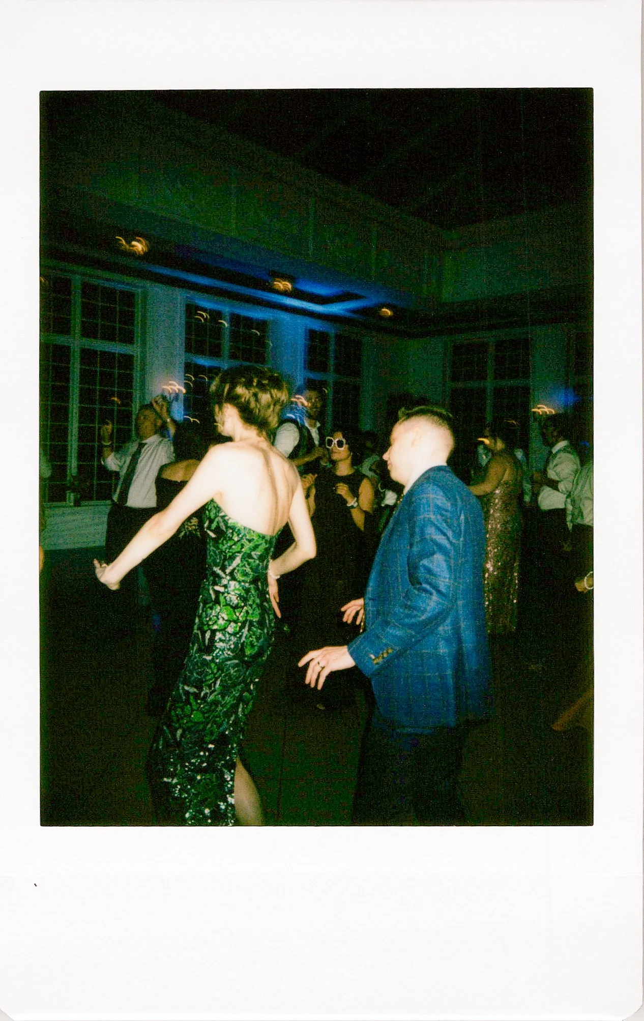 People dancing at a social event in a room with large windows and blue lighting.