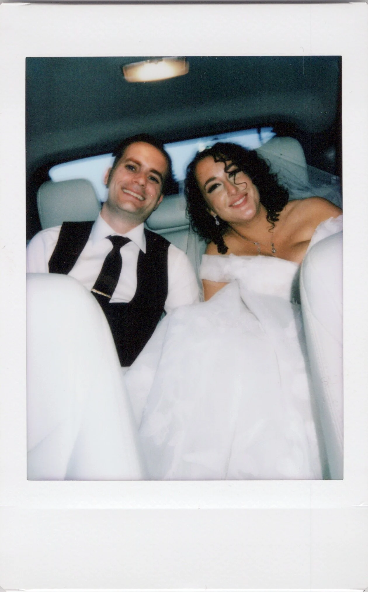 A newlywed couple sitting in the backseat of a vehicle, smiling for a photograph. The groom is dressed in a black vest, white shirt, and black tie, while the bride is in a white wedding gown with off-the-shoulder sleeves and jewelry.
