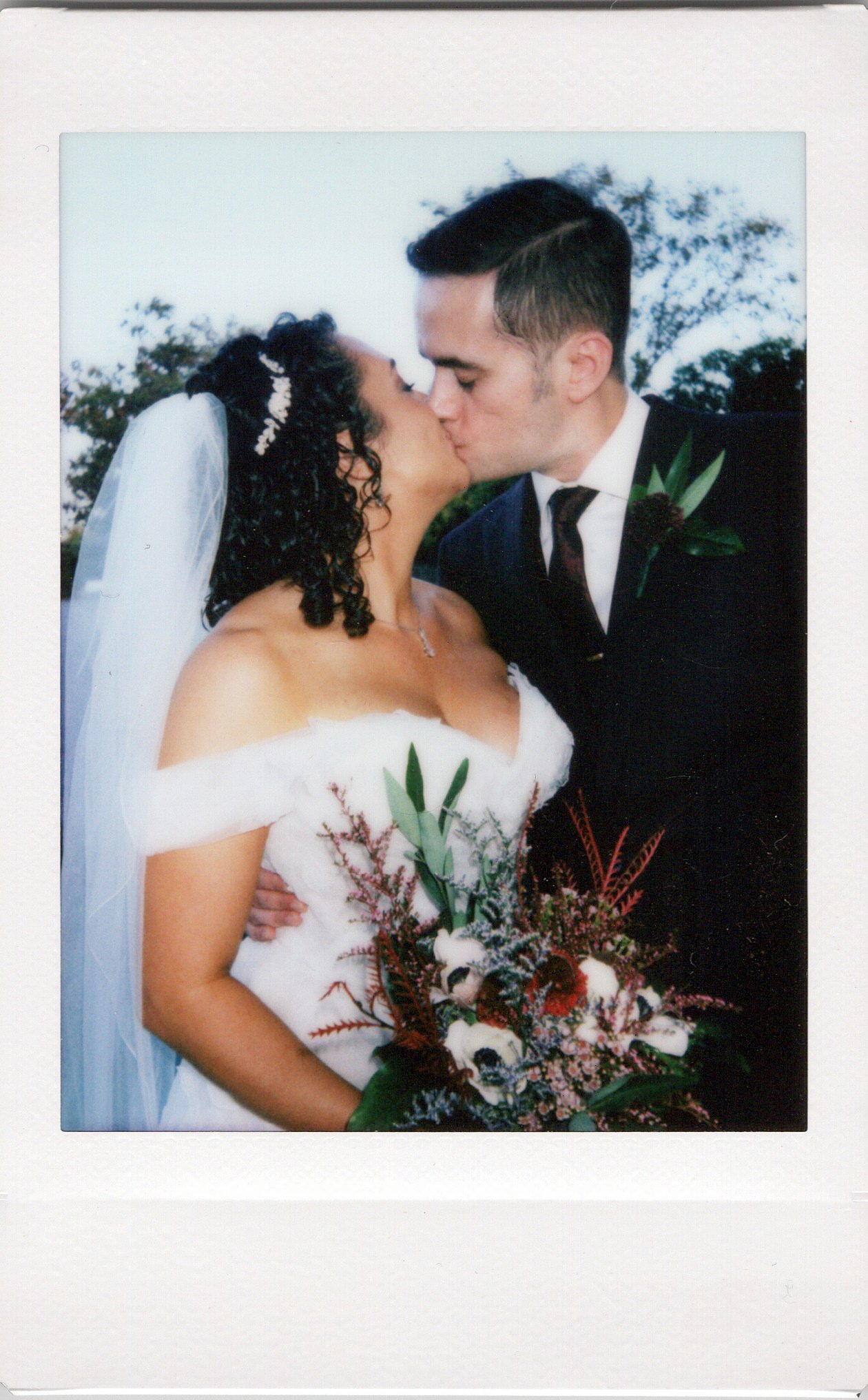 A bride and groom sharing a kiss on their wedding day, with the bride holding a bouquet of flowers, outdoors with trees in the background. Chicago polaroid photo booth