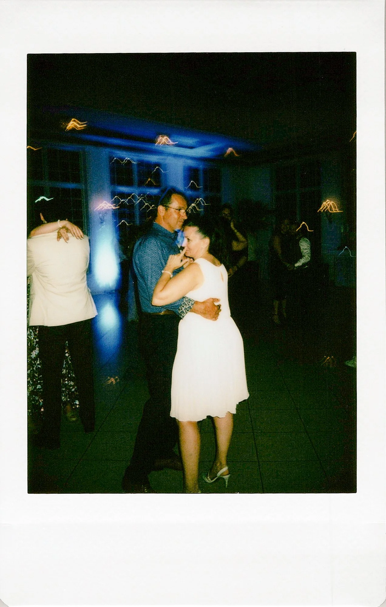 Couple dancing closely at a formal event during nighttime, surrounded by other guests in a dimly lit room with blue lighting and windows in the background.