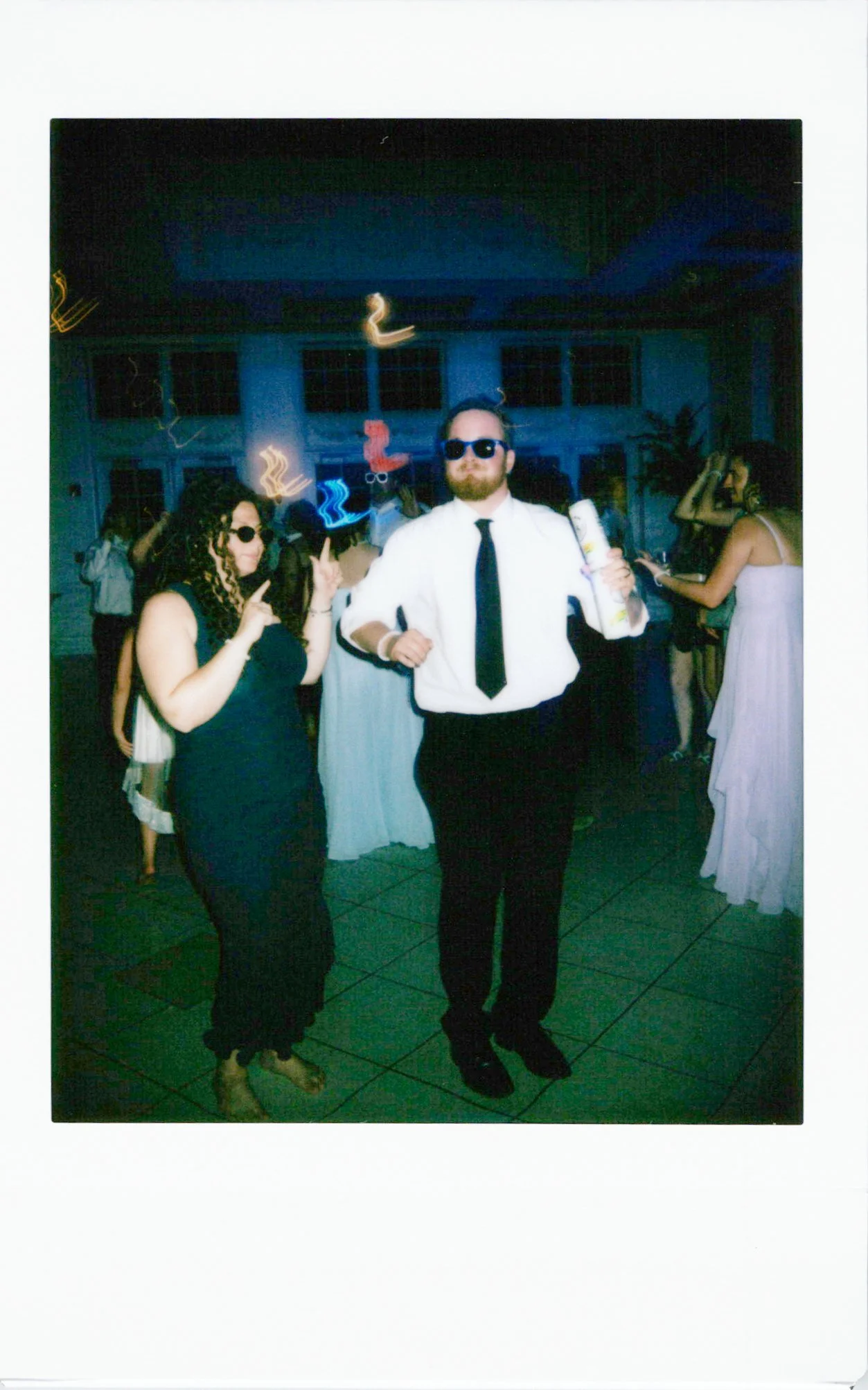 A man in a white shirt, black tie, and sunglasses holding a drink at a party. A woman in black dress and sunglasses standing nearby. Several people in the background at a dimly lit party or dance event.