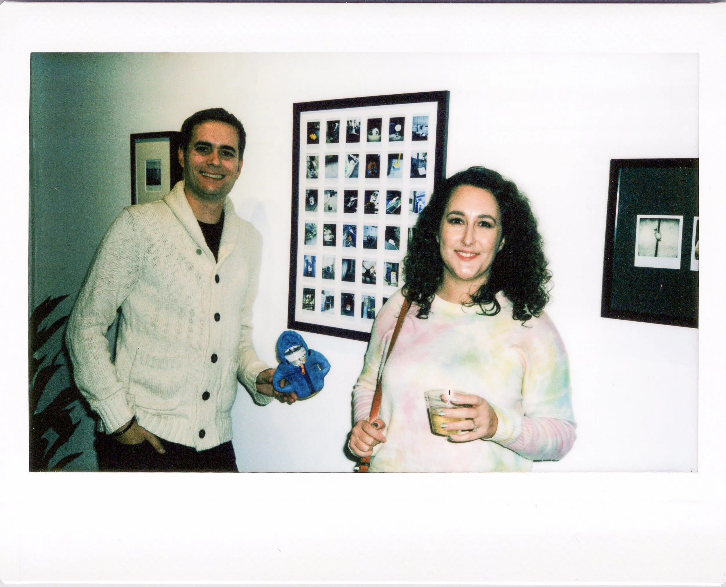 A man and a woman are standing indoors near a wall with framed pictures. The man is wearing a cream-colored button-up sweater, smiling, and holding a small plush toy. The woman has curly hair, is smiling, holding a drink, and wearing a pastel-colored tie-dye sweater.