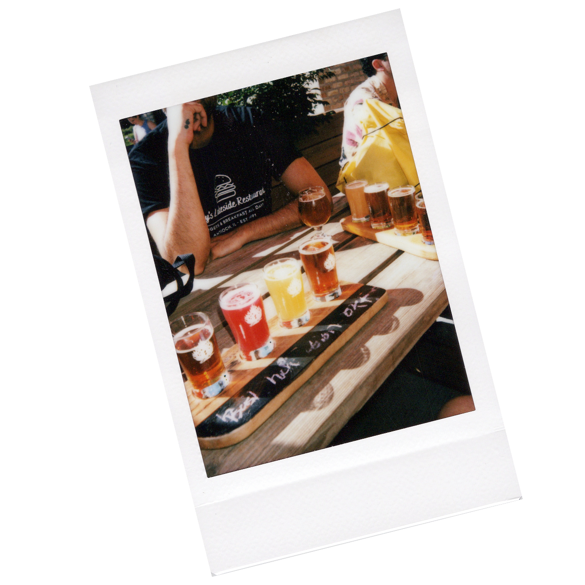 A person sitting at a wooden table with a tray of various colored beers in small glasses, outdoors in a garden setting.