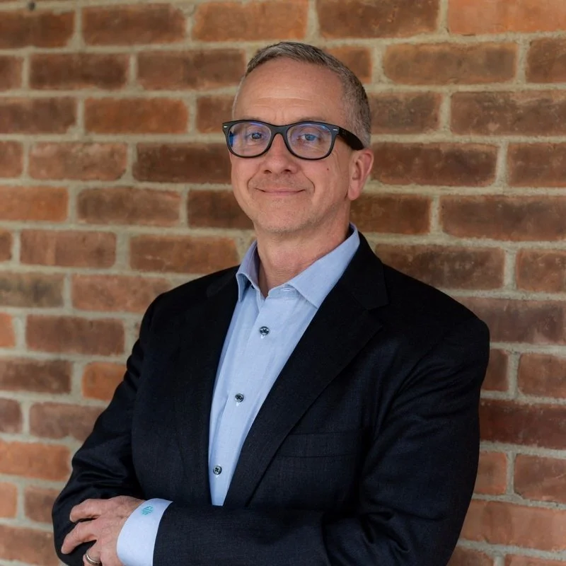 A man with short hair, wearing glasses, a light blue dress shirt, and a dark blazer, standing with arms crossed in front of a brick wall, smiling at the camera; headshot of Board Chair Chris DeRuyver