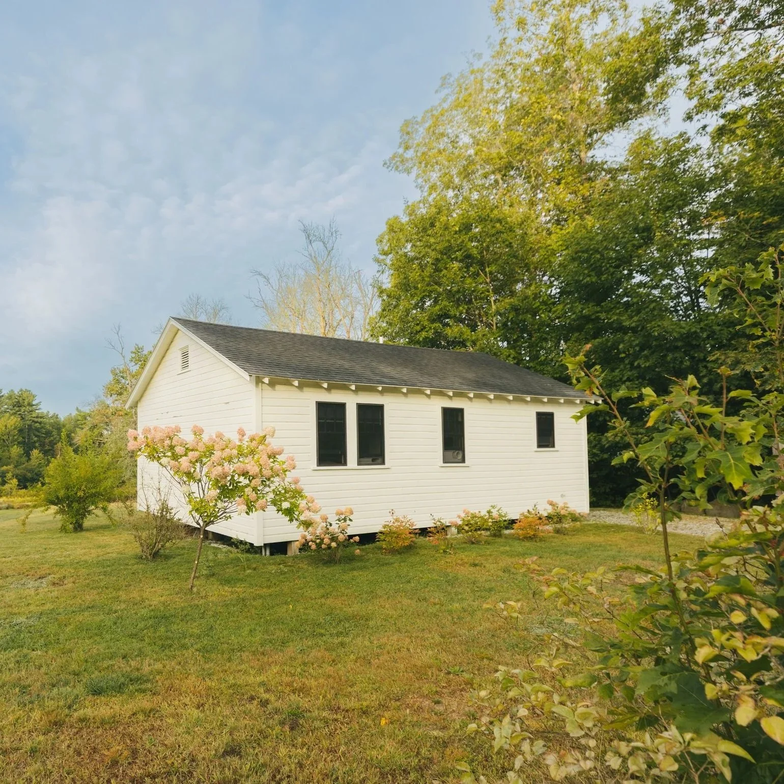A white house with black windows surrounded by green grass, bushes, and trees on a sunny day with blue sky.