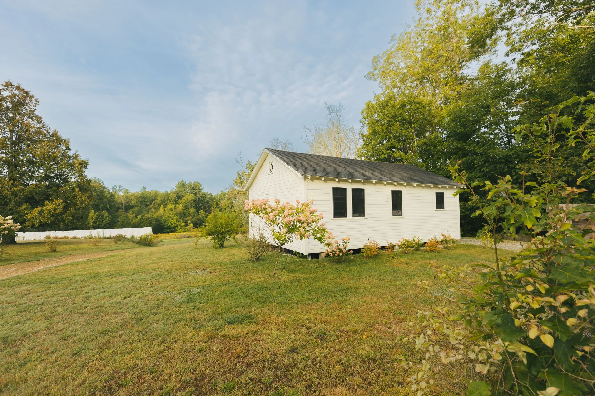 A white house with a black roof surrounded by green trees and a garden with flowers, under a partly cloudy sky.