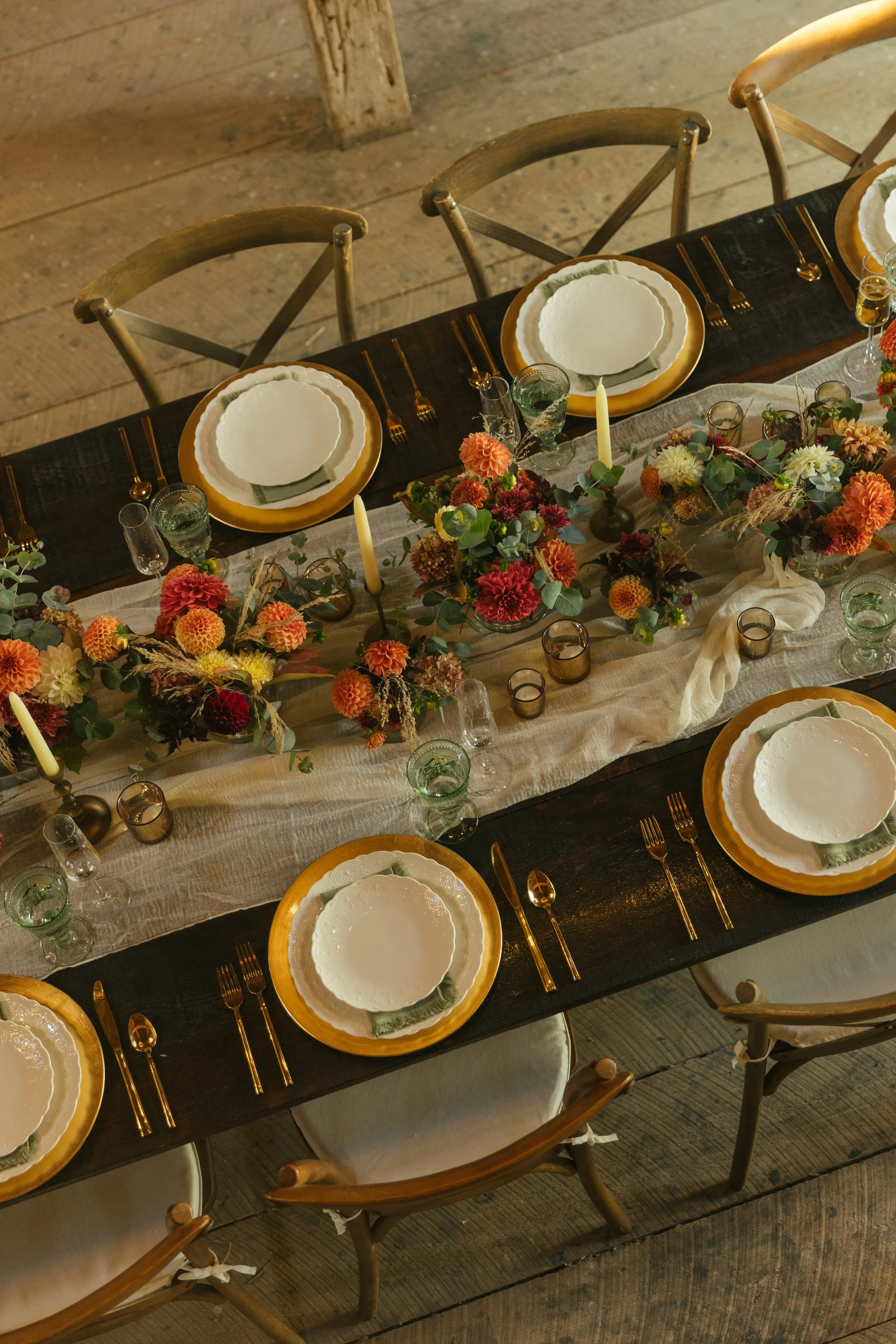 A dining table set for a formal event with white plates, green napkins, gold utensils, candles, and floral centerpieces with orange, red, yellow, and white flowers.