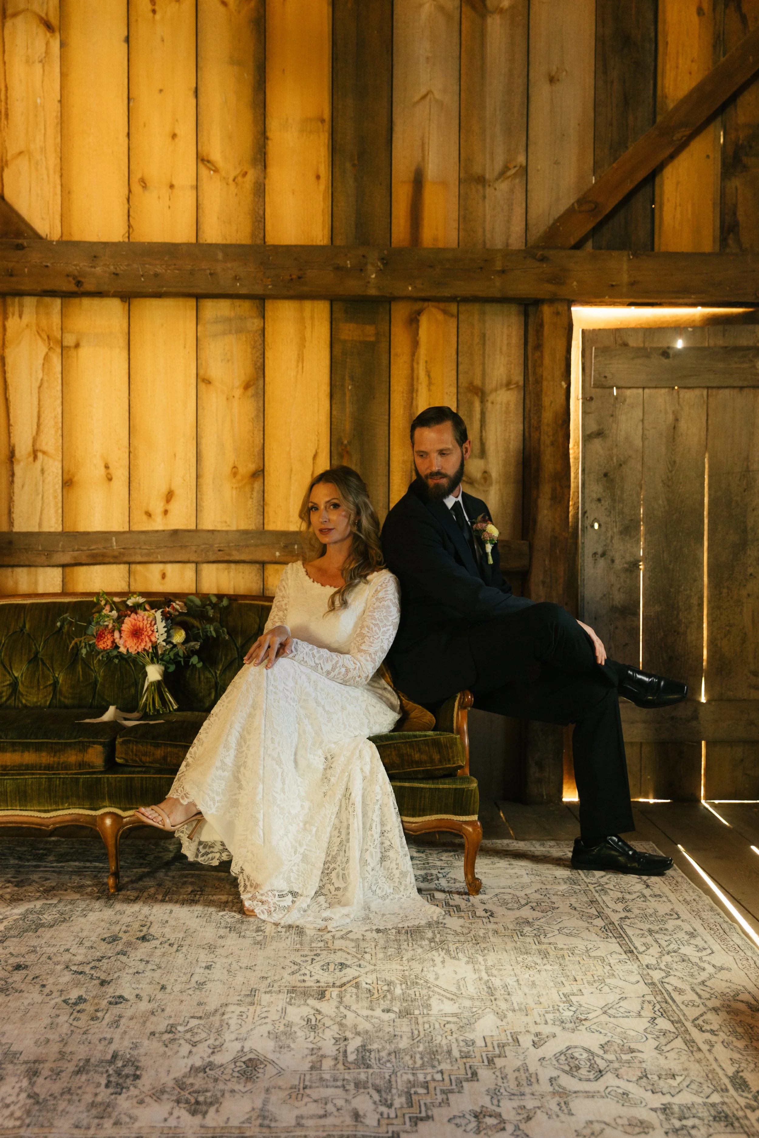 A bride in a white lace wedding dress and a groom in a black suit sit back-to-back on a vintage green velvet sofa inside a rustic wooden barn with warm lighting.
