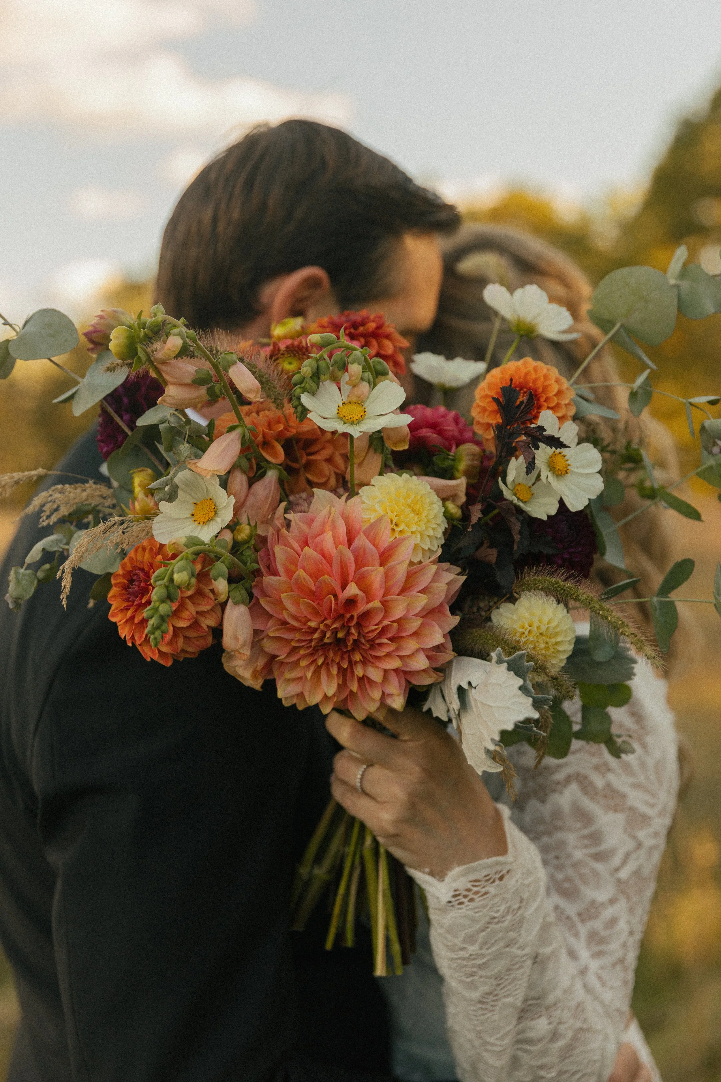A couple hugging and holding a large bouquet of colorful flowers outdoors.