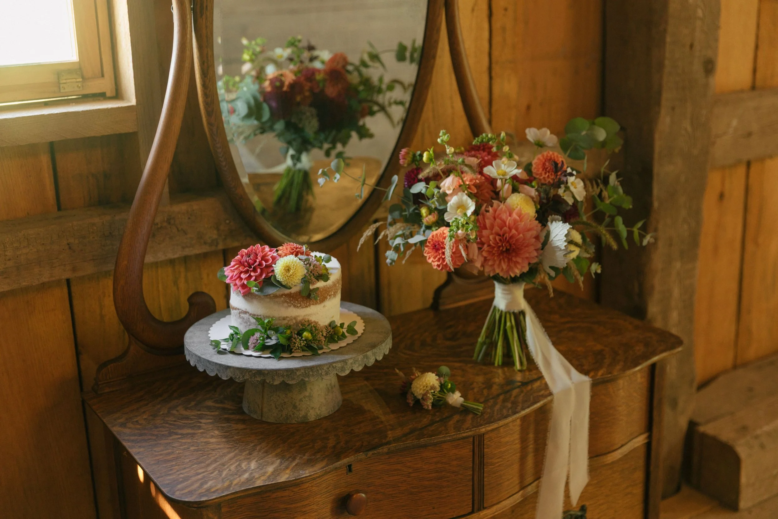 A small cake decorated with pink and yellow flowers, a bouquet of pink, white, and green flowers in a vase, and a mirror reflecting the bouquet, all placed on a wooden dresser against wooden walls.