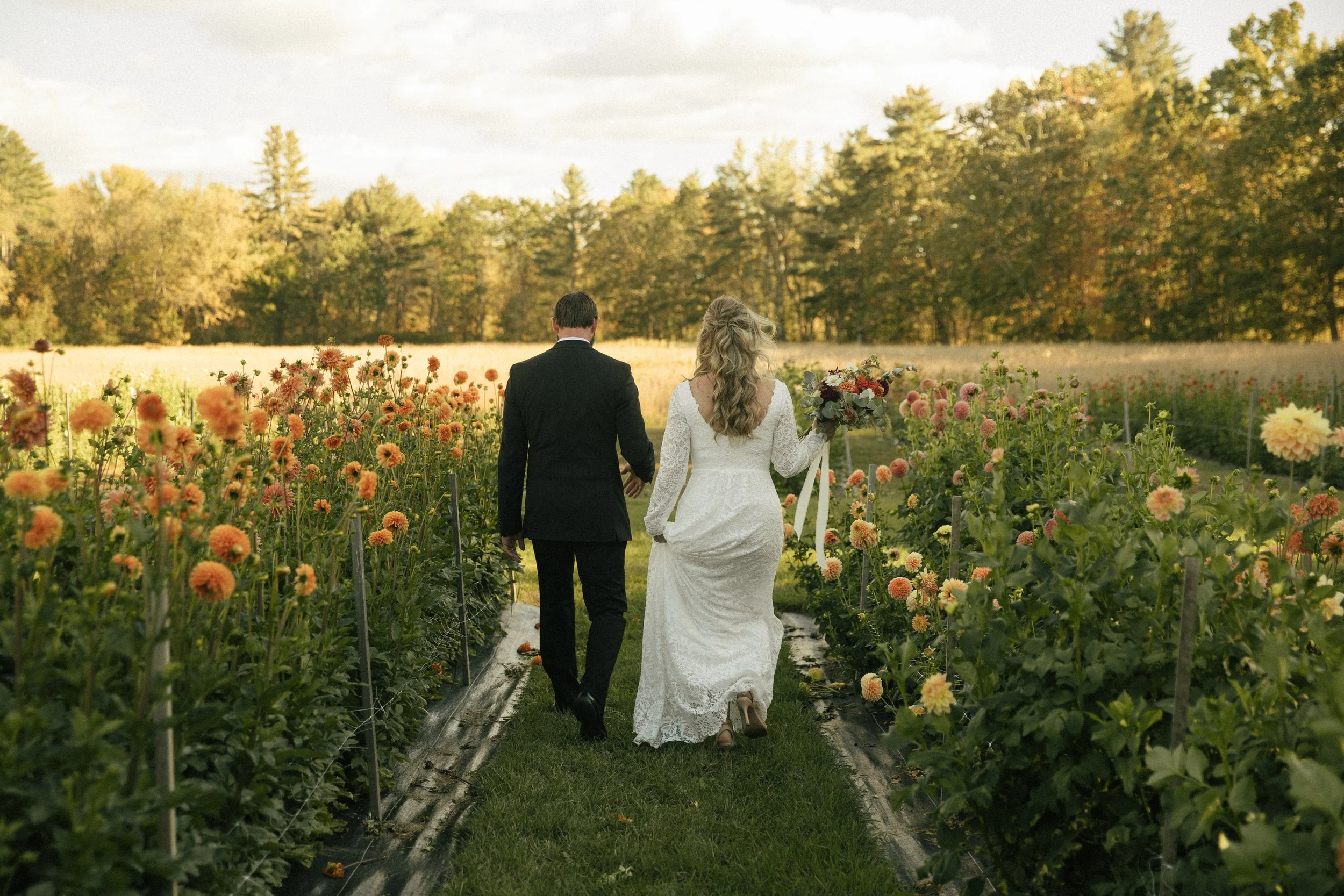A newlywed couple walking hand in hand through a flower field during sunset, with a woman in a white lace dress holding a bouquet and a man in a black suit.