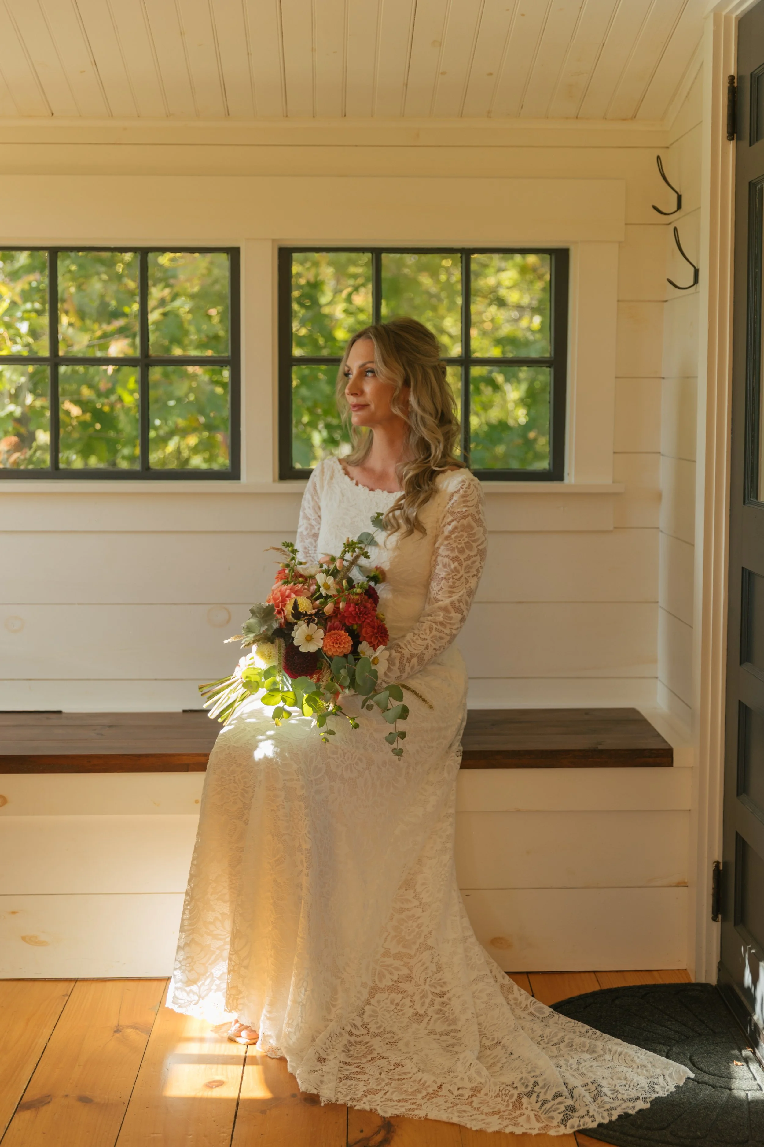 A woman in a white lace dress holding a bouquet of flowers, sitting indoors near windows with a view of greenery outside.