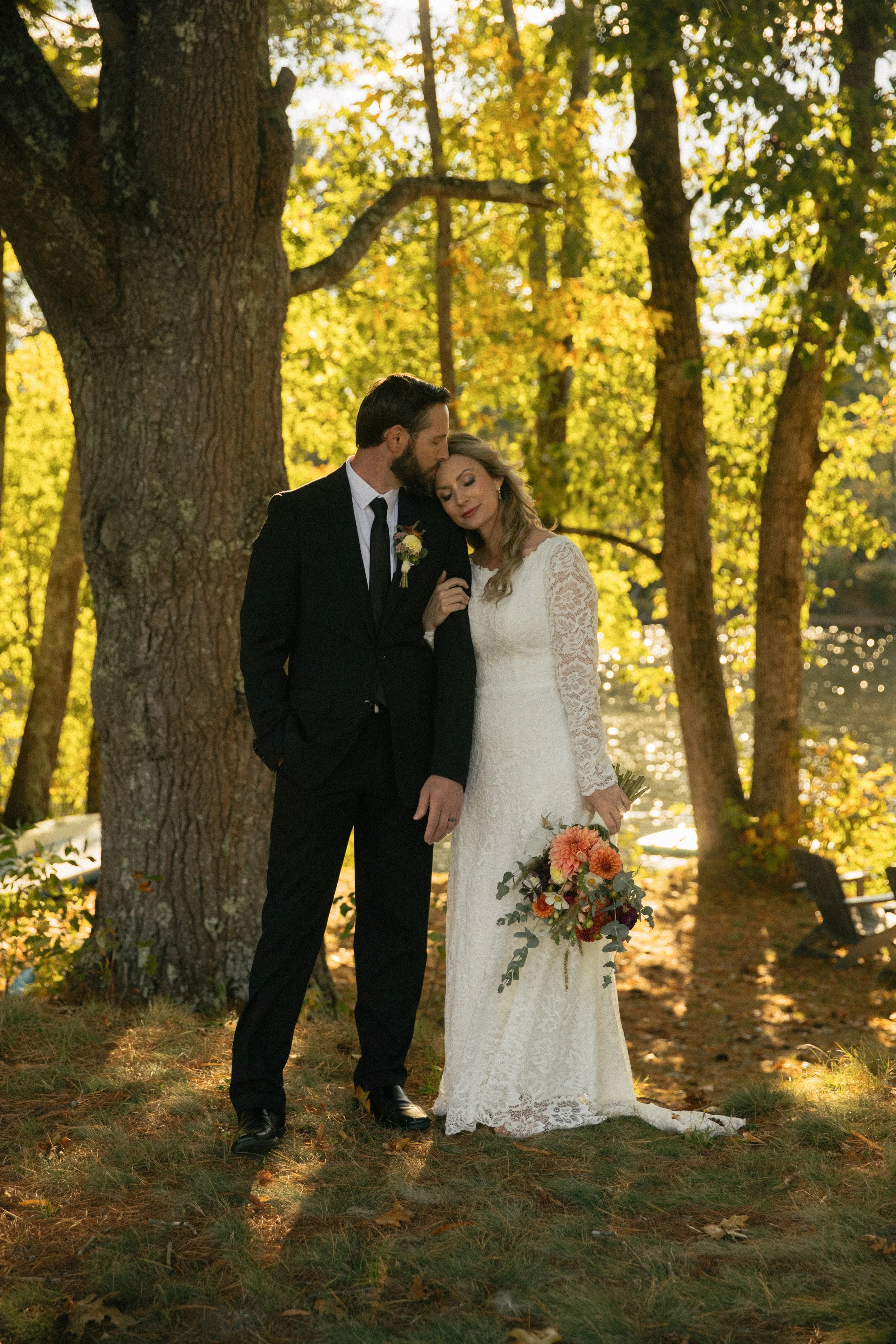 A bride and groom standing together outdoors in a forested area during autumn, with the groom kissing the bride's forehead. The bride is holding a bouquet of flowers, and both are dressed in wedding attire.