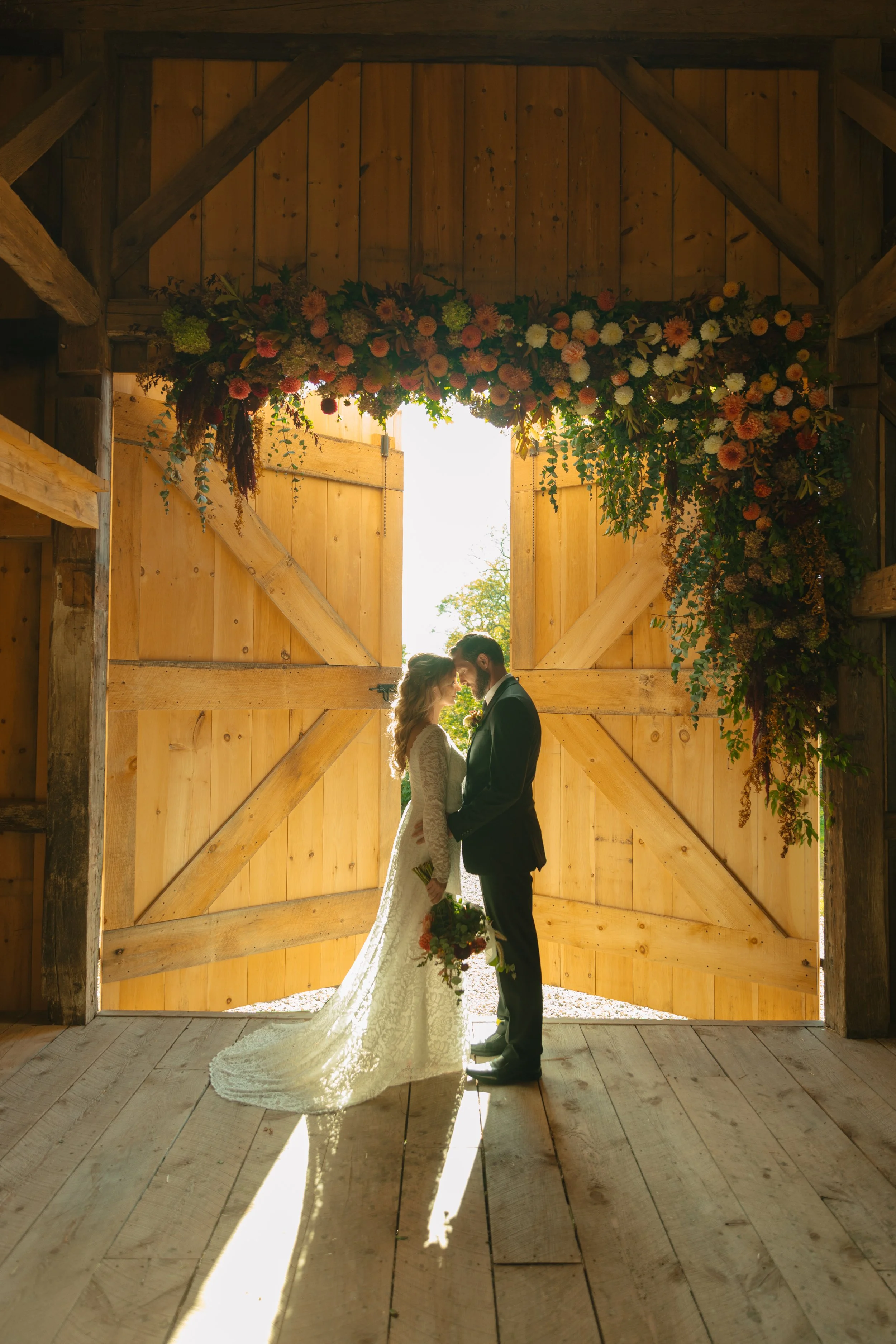 A bride and groom standing close together in front of open barn doors decorated with a floral arrangement, backlit by sunlight, creating a romantic silhouette.