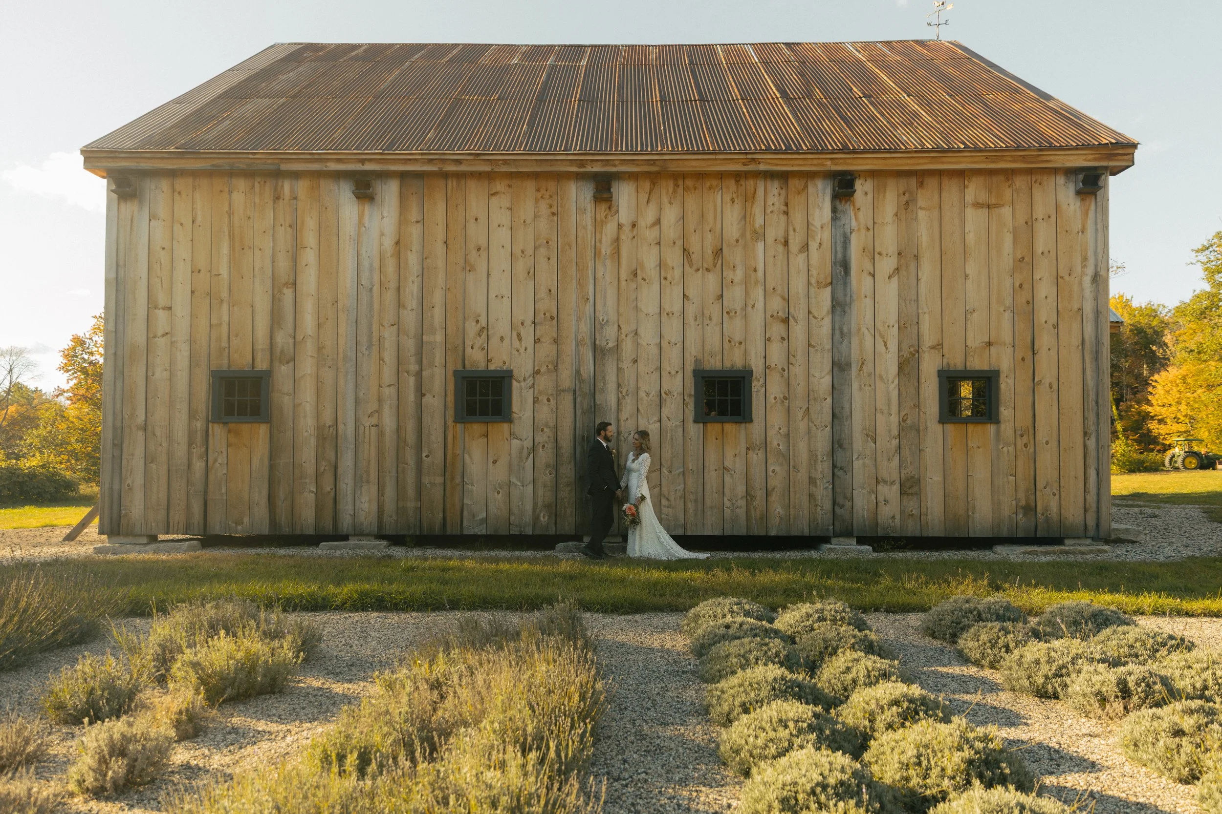 A bride and groom in wedding attire holding hands and facing each other in front of a rustic barn with wooden exterior and small windows. The scene is outdoors during daytime with clear sky and surrounding landscape.