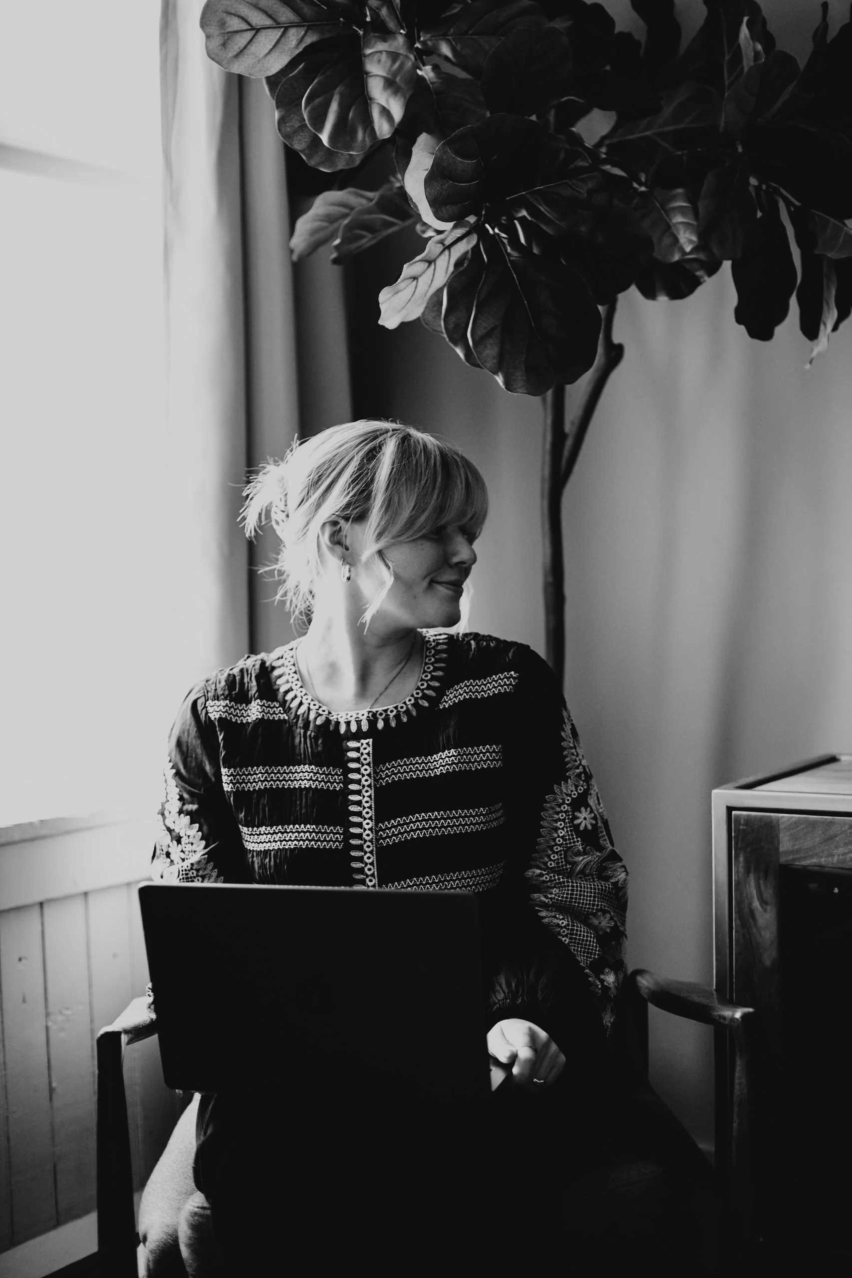 A woman with shoulder-length hair sitting next to a laptop, looking to her left, in a room with a large indoor plant and a window with curtains, in black and white.