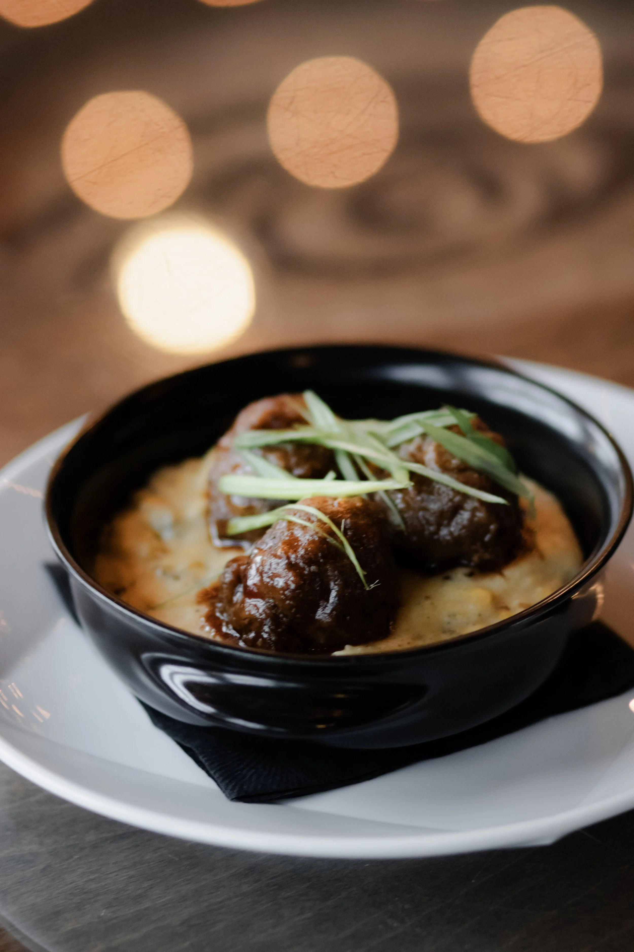 Meatballs in sauce with green garnish, served in a black bowl on white plate with a black napkin underneath.