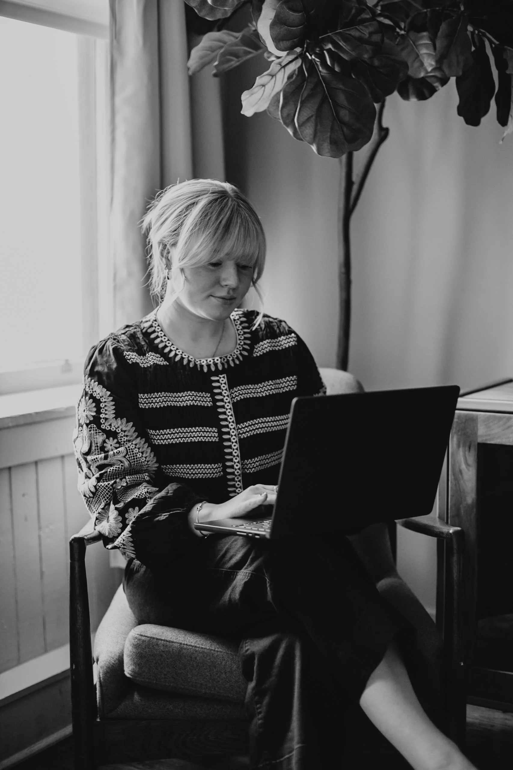 A woman sitting on a chair by a window, working on a laptop, with a large plant behind her.