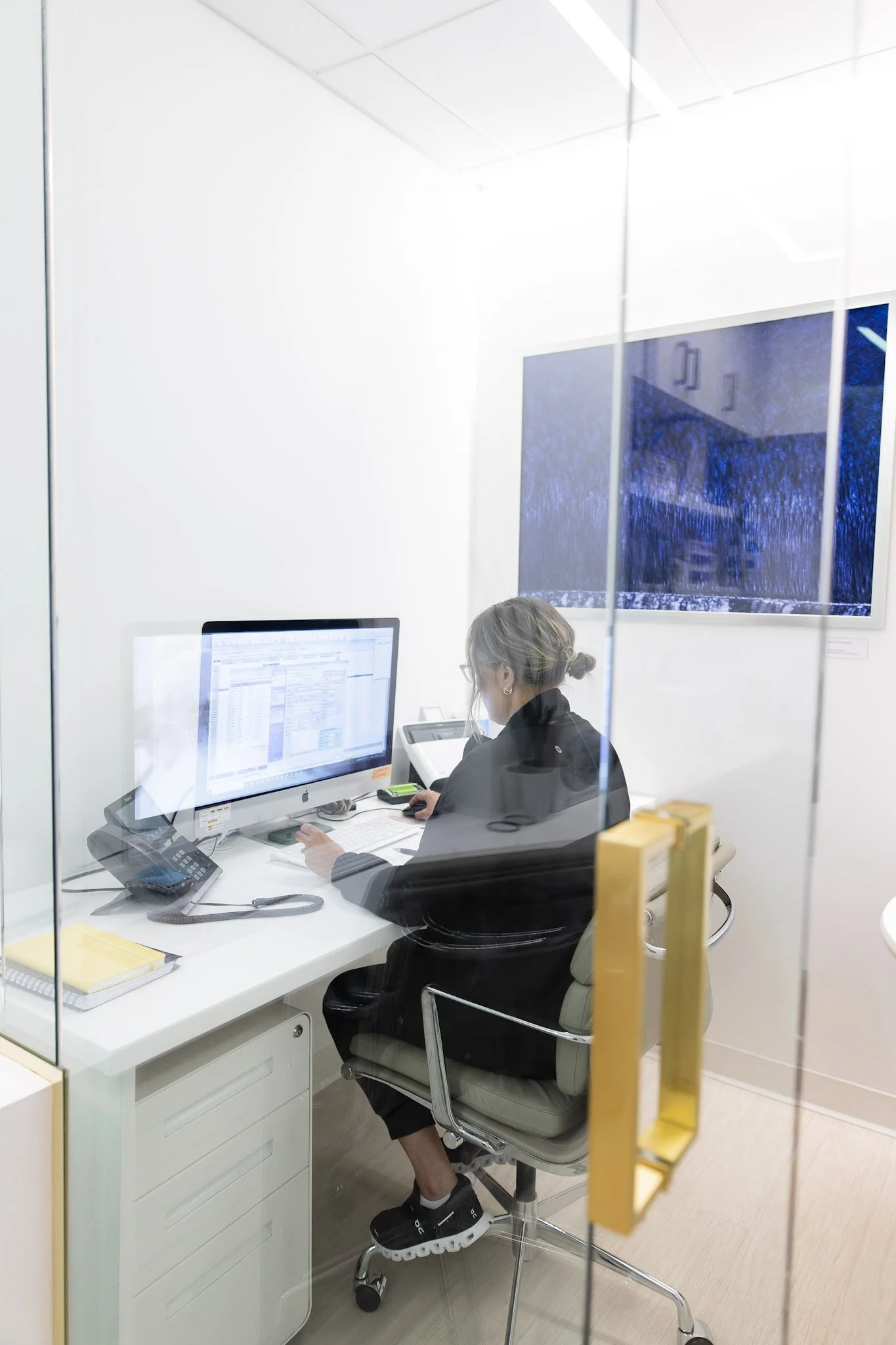 A woman working at her desk in a modern office, seen through a glass wall. She has gray hair tied back, is wearing glasses and a black jacket, and is using a desktop computer and a tablet. A monitor displaying a blue screen is on the wall beside her.