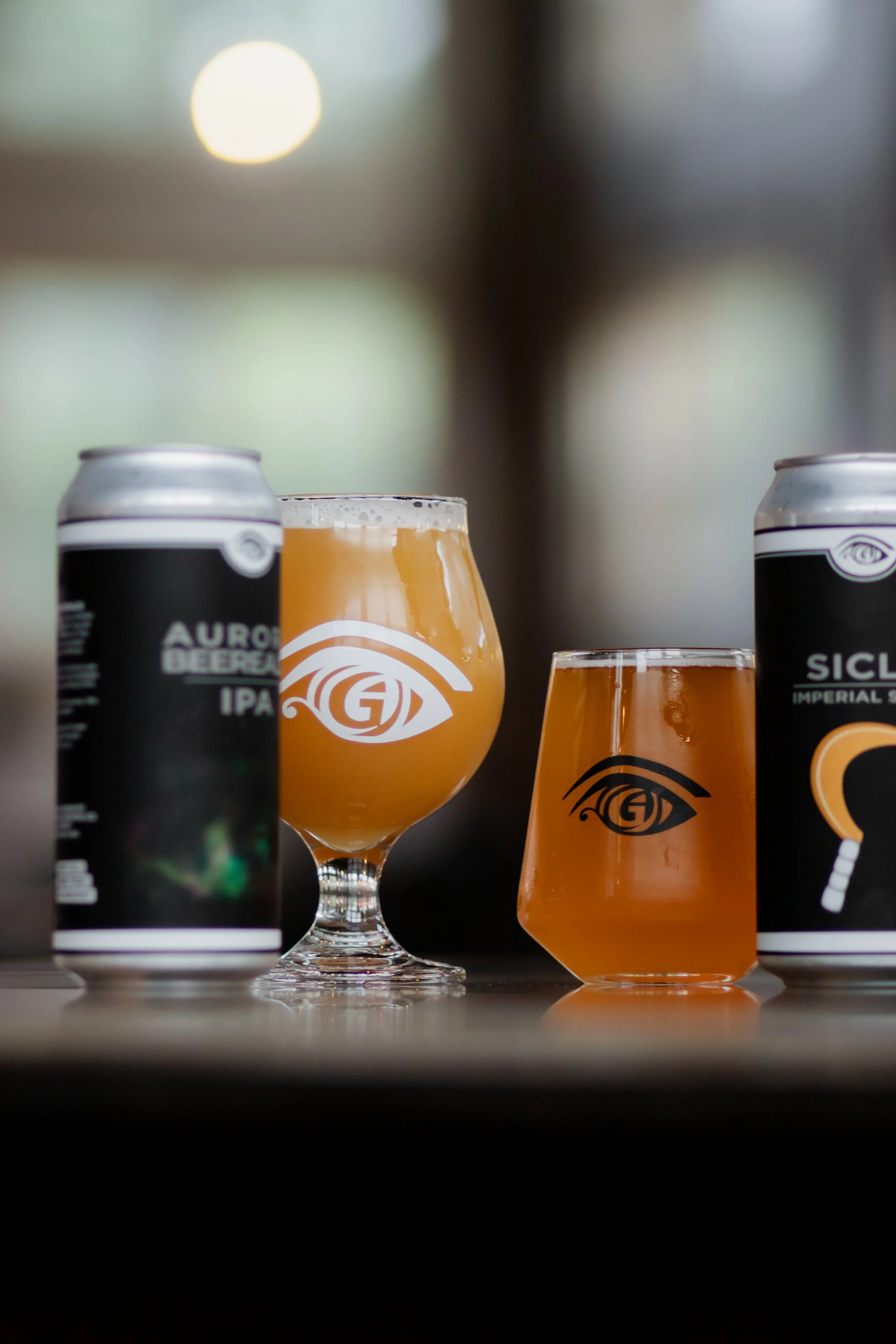 Two cans of beer and two glasses of beer with logos, on a dark surface.