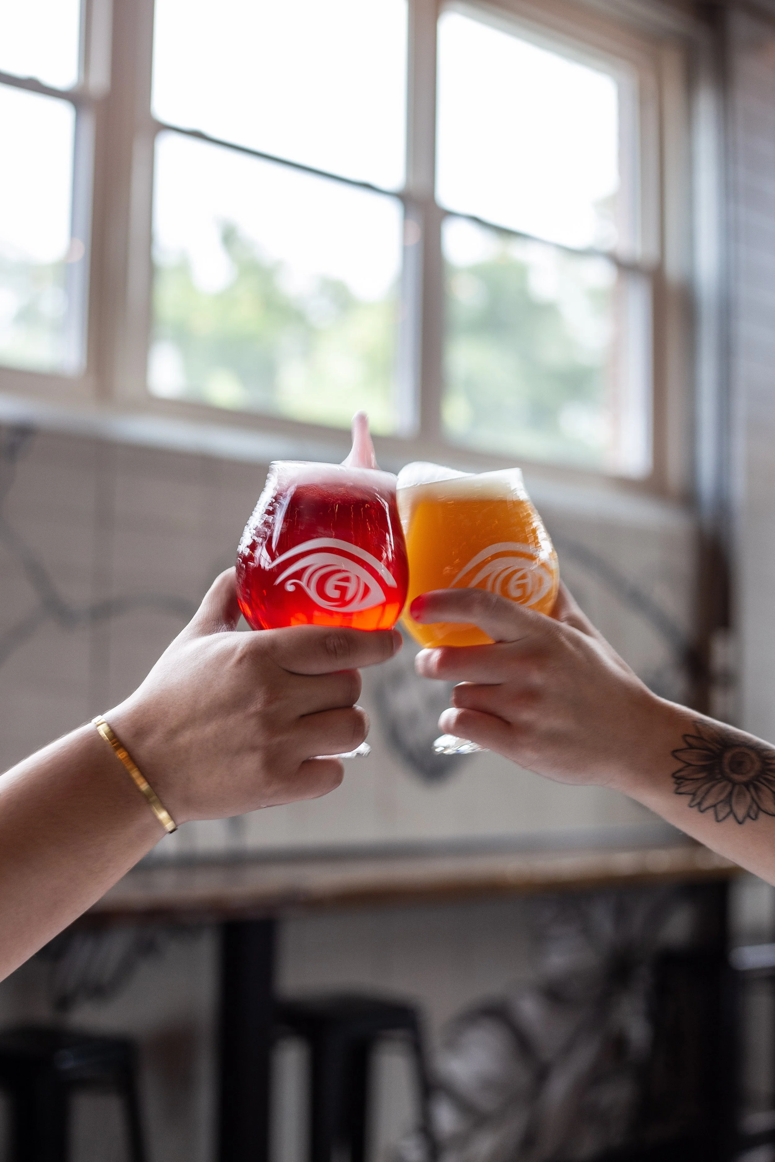 Two hands holding glasses of colorful beer, one red and one orange, clinking together inside a room with large windows and a blurred background.