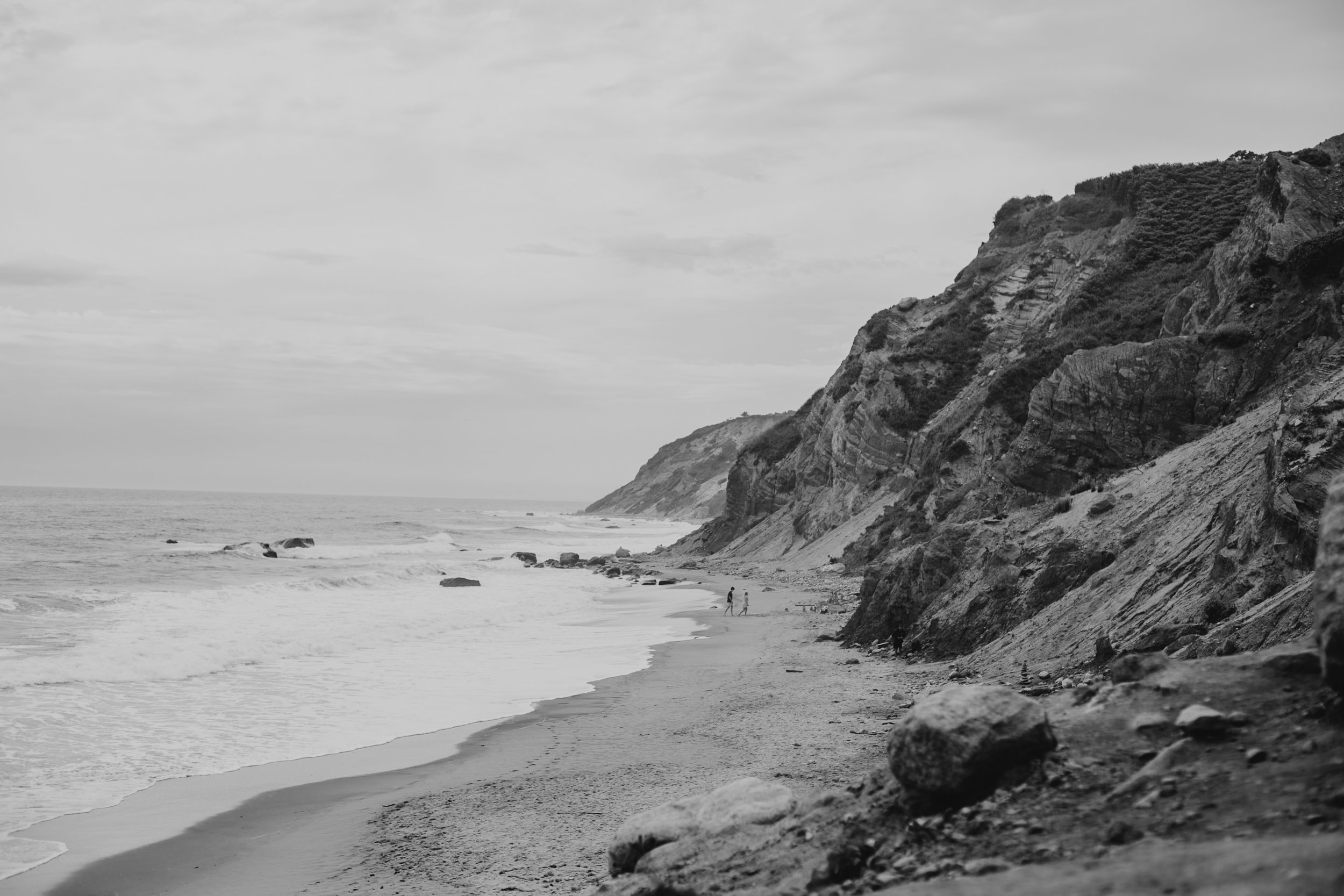Black-and-white photo of a beach with cliffs on the right side, a few people walking along the shoreline, and rocks scattered across the sand.