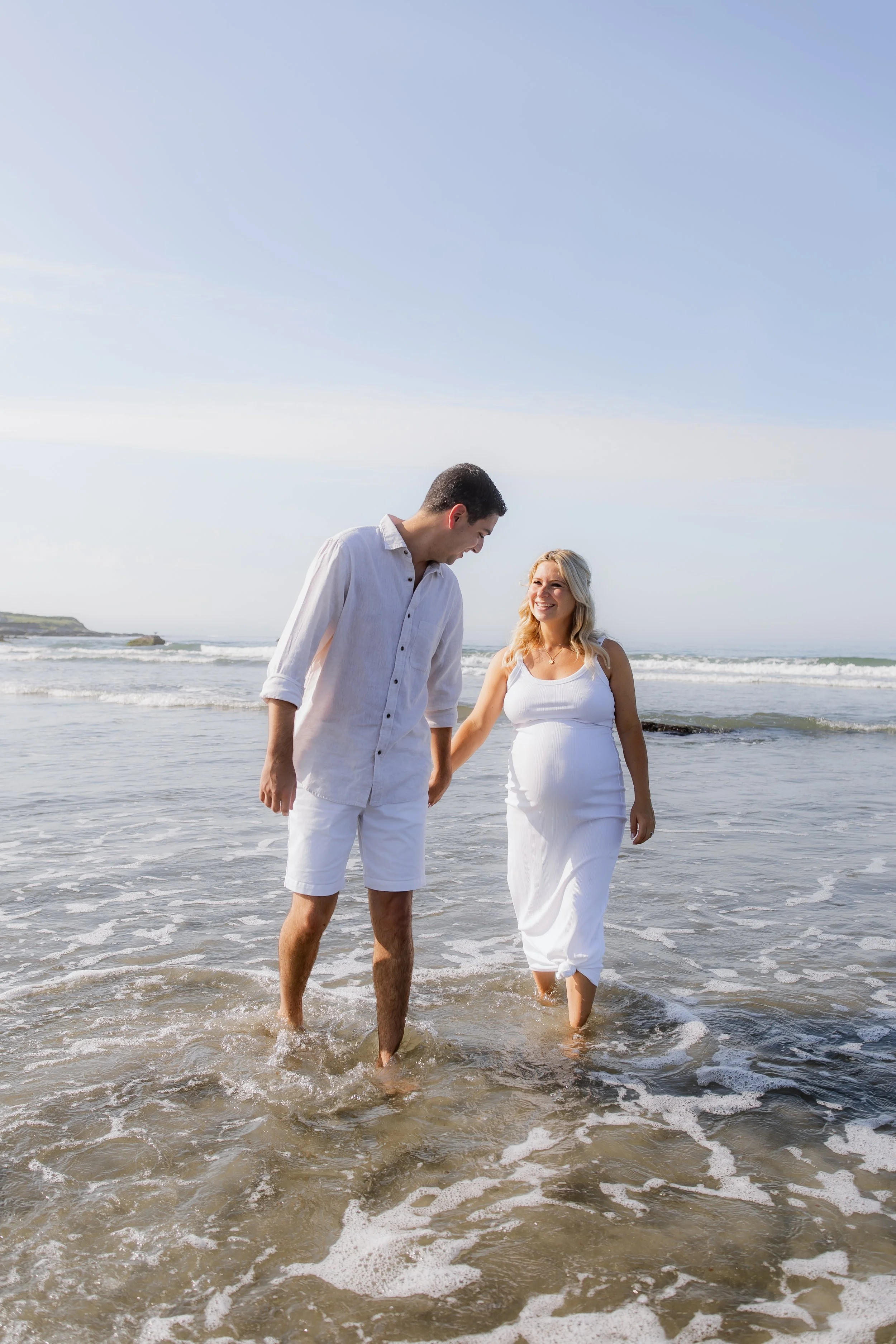 A pregnant woman and a man, possibly her partner, walking hand in hand in the ocean water at the beach, both dressed in white, with waves and a blue sky in the background.