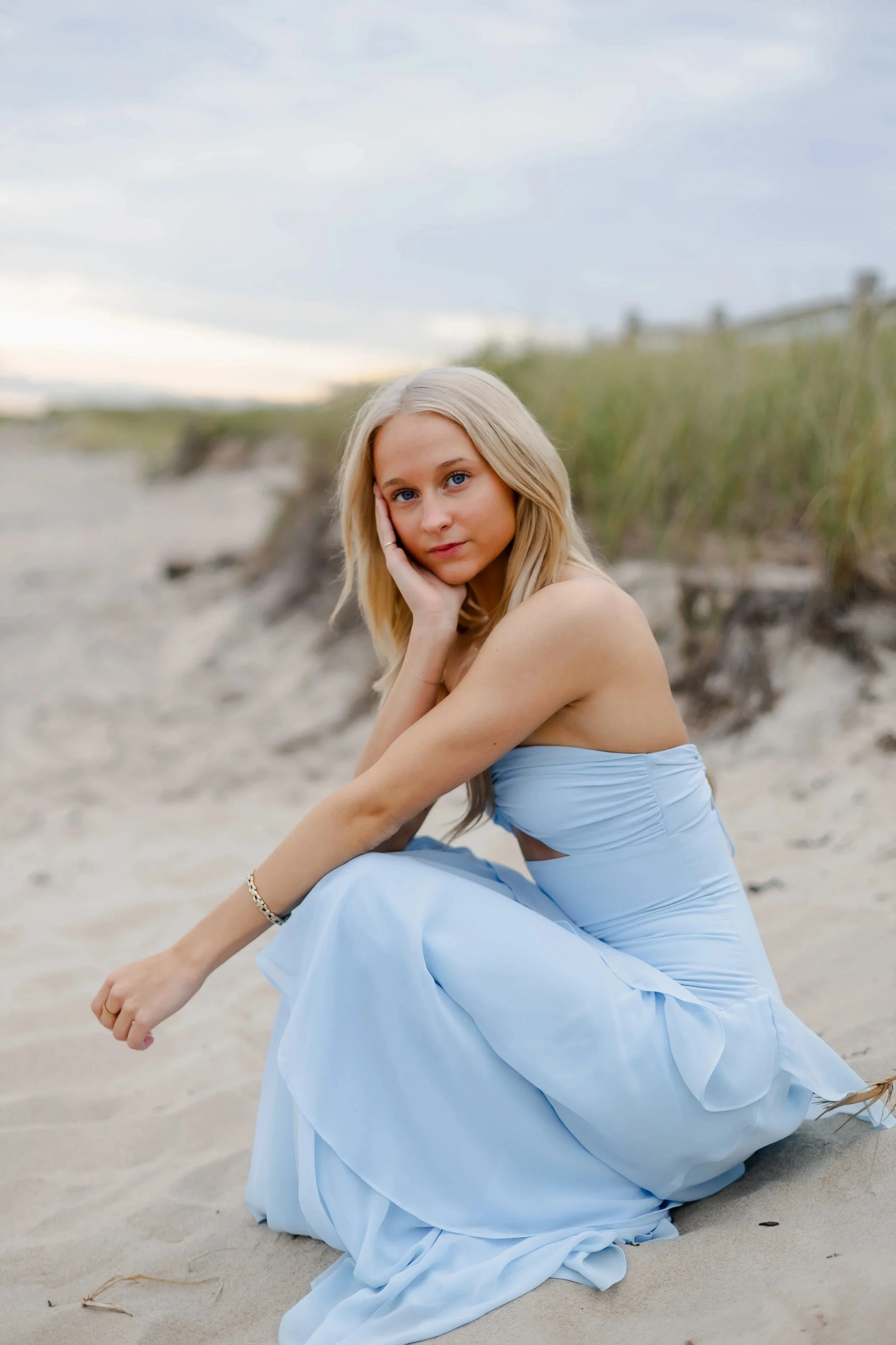 A woman with blonde hair wearing a light blue dress sitting on the sand at a beach, with grassy dunes and cloudy sky in the background.