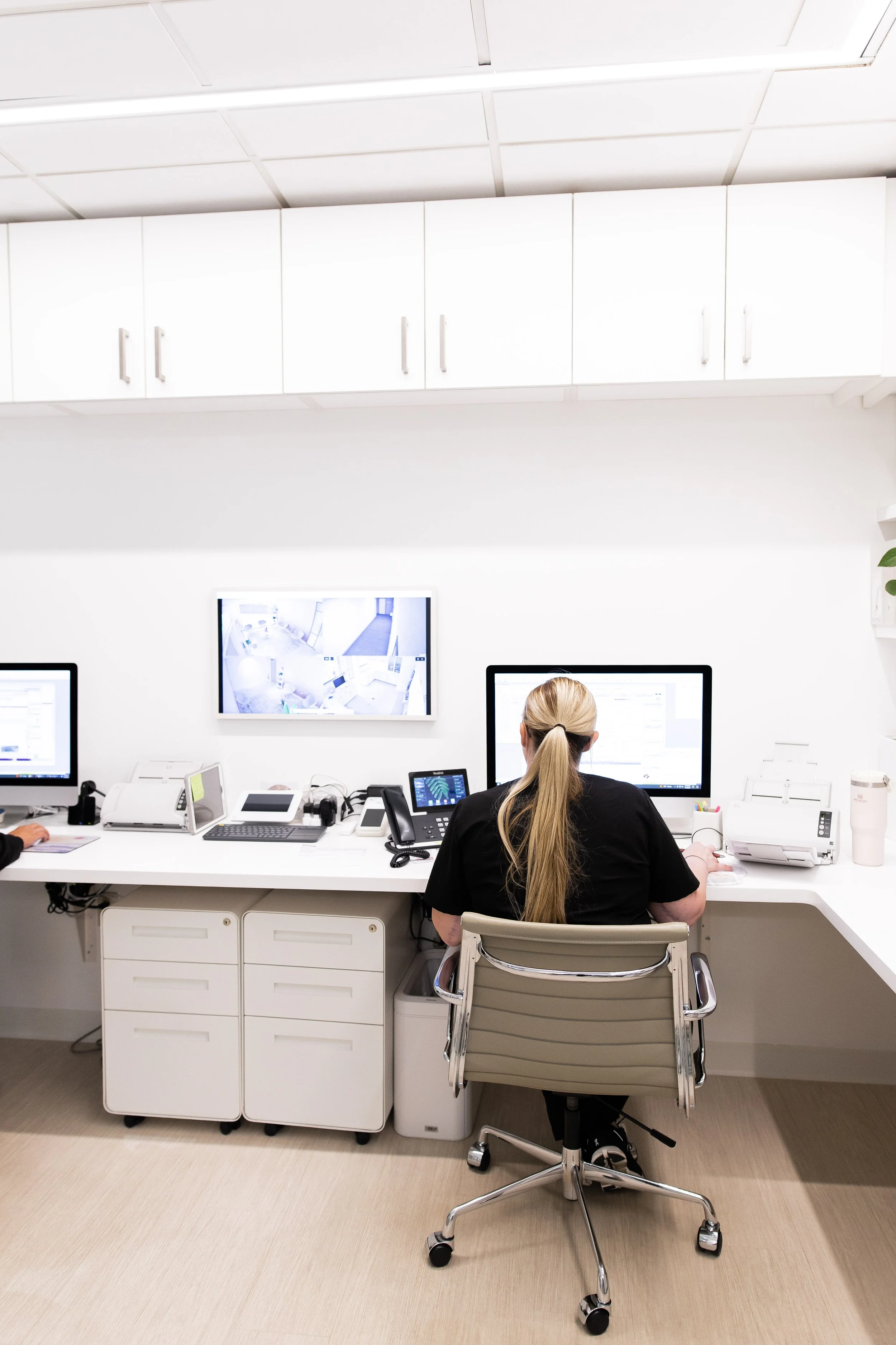 Healthcare professional sitting at a white desk in front of computer monitors, with office equipment and cabinets in a clean, modern office space.