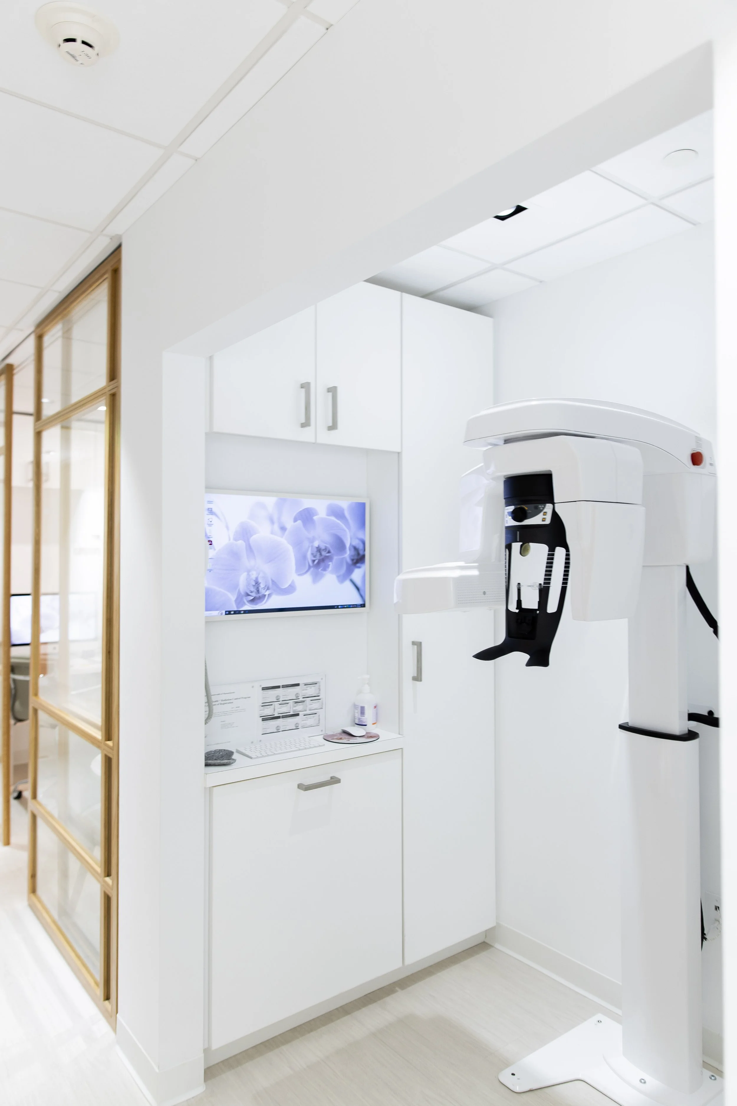 Dental X-ray machine in a bright, white dental clinic room with cabinets, a monitor displaying purple orchids, and a partially visible wooden framed glass door.