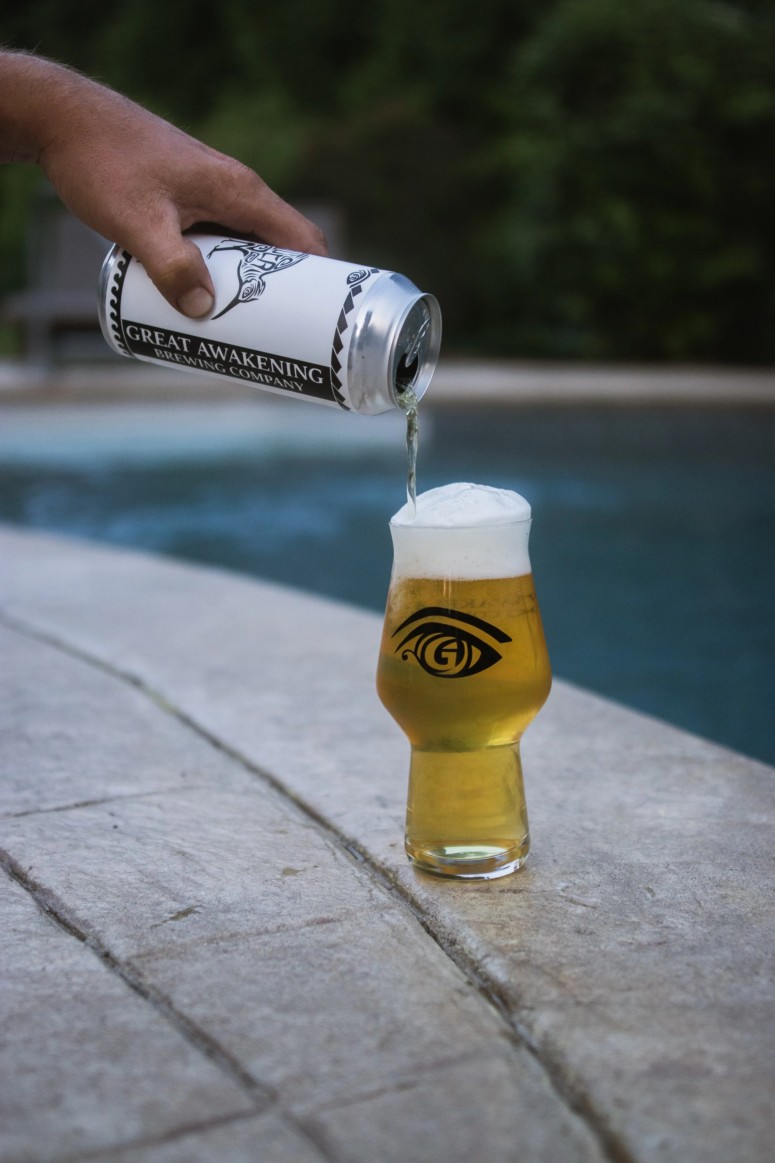 A hand pouring beer from a can labeled 'Great Awakening Brewing Company' into a glass with a 'Great Awakening' eye logo, set on a poolside stone surface.