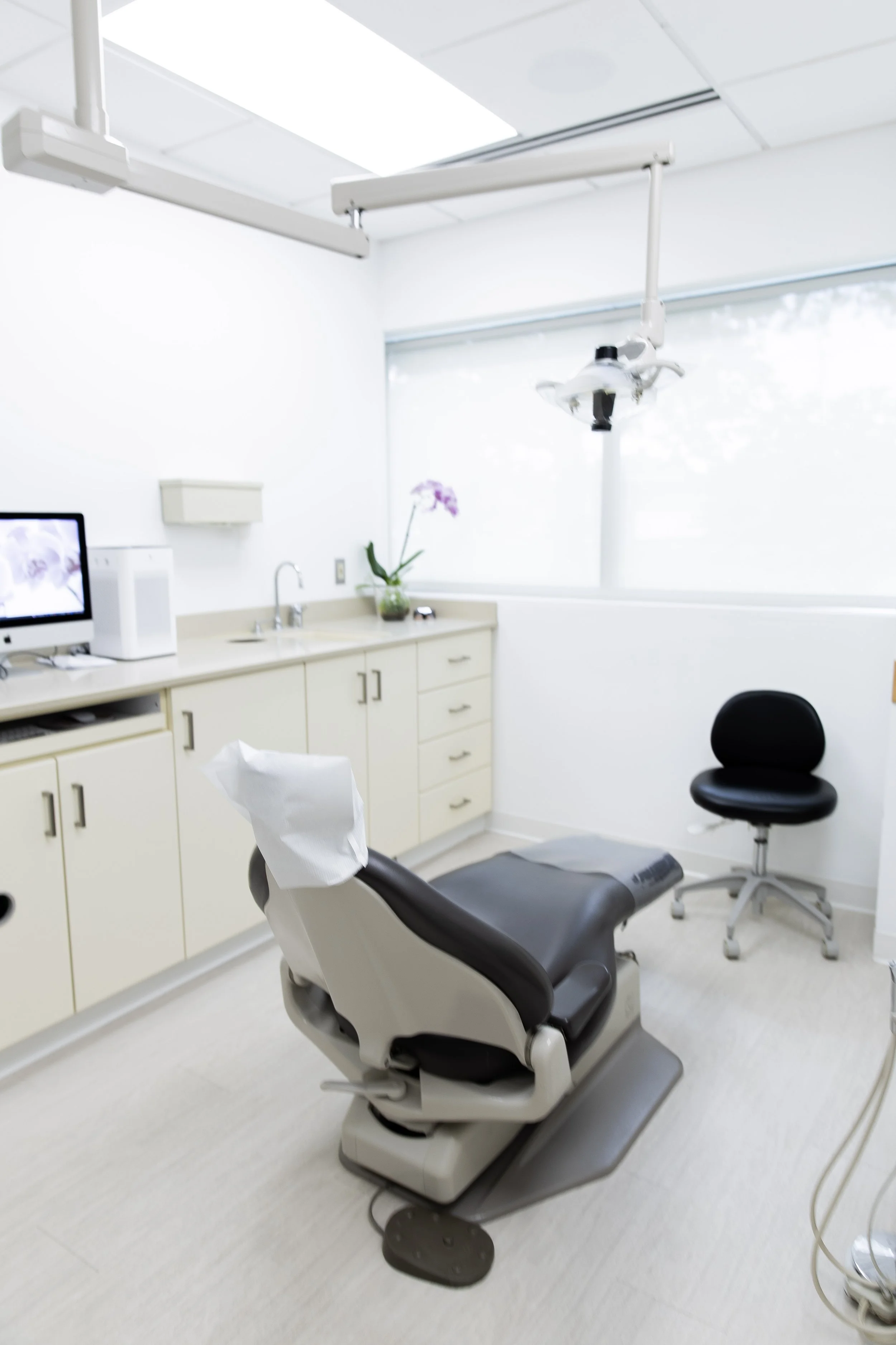 A clean dental examination room with a dentist chair, a computer monitor, a small cabinet, a sink, a chair, and dental equipment, with a large window allowing natural light.