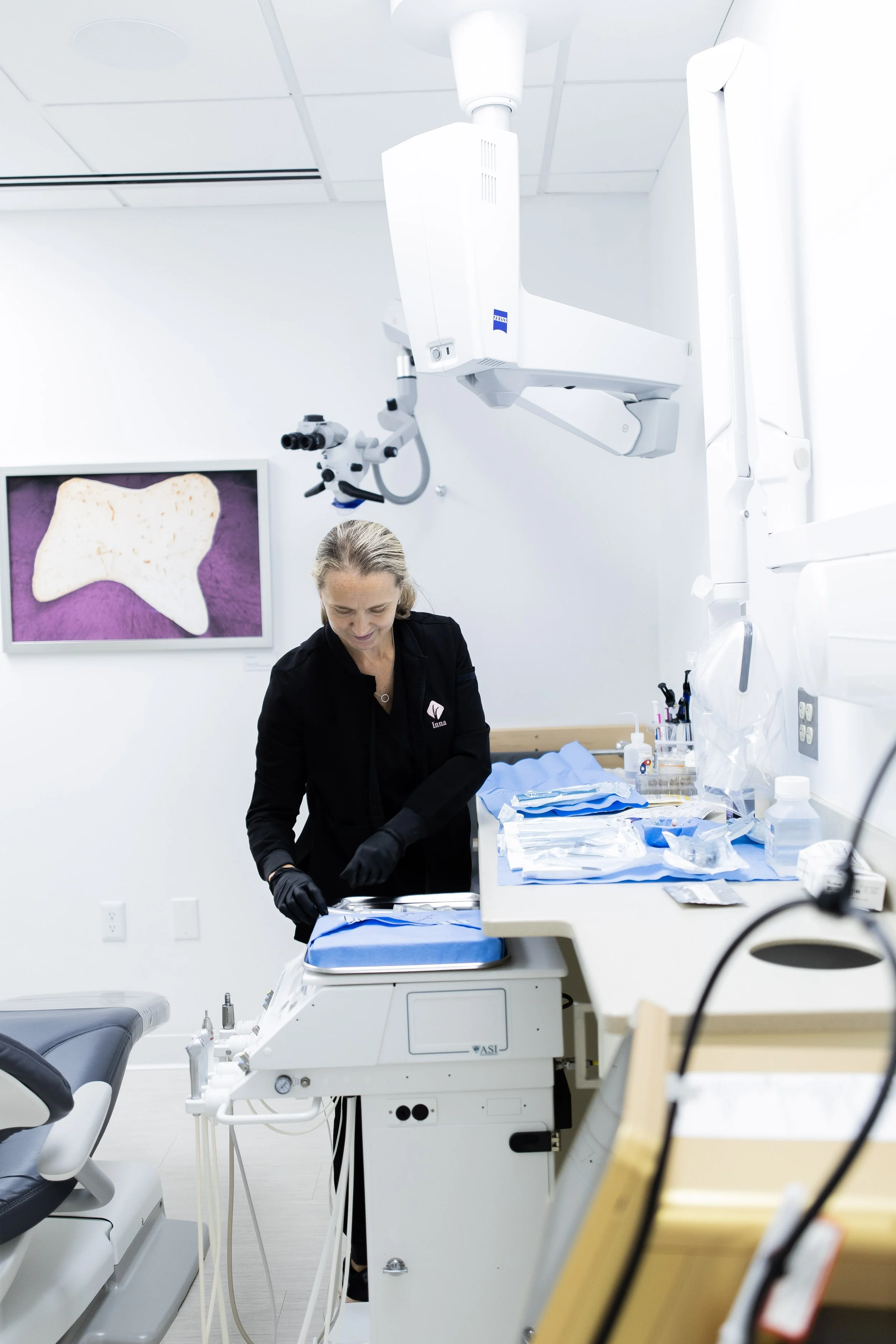 A dentist wearing black gloves preparing dental instruments in a modern dental clinic, with dental equipment and a monitor displaying a dental scan or X-ray.