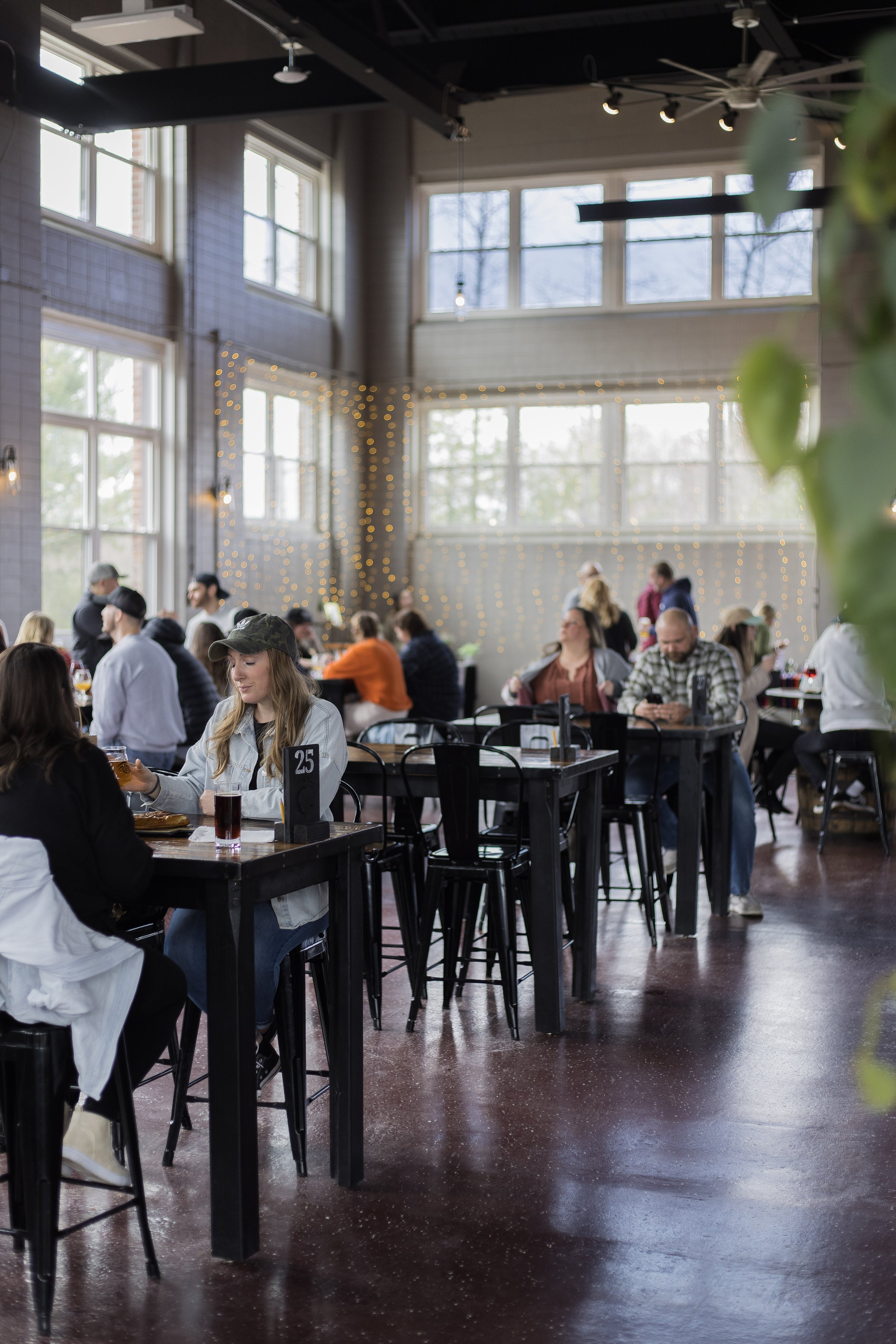People sitting and socializing at tables in a spacious, well-lit cafe or restaurant with large windows, string lights, and dark wooden floors.
