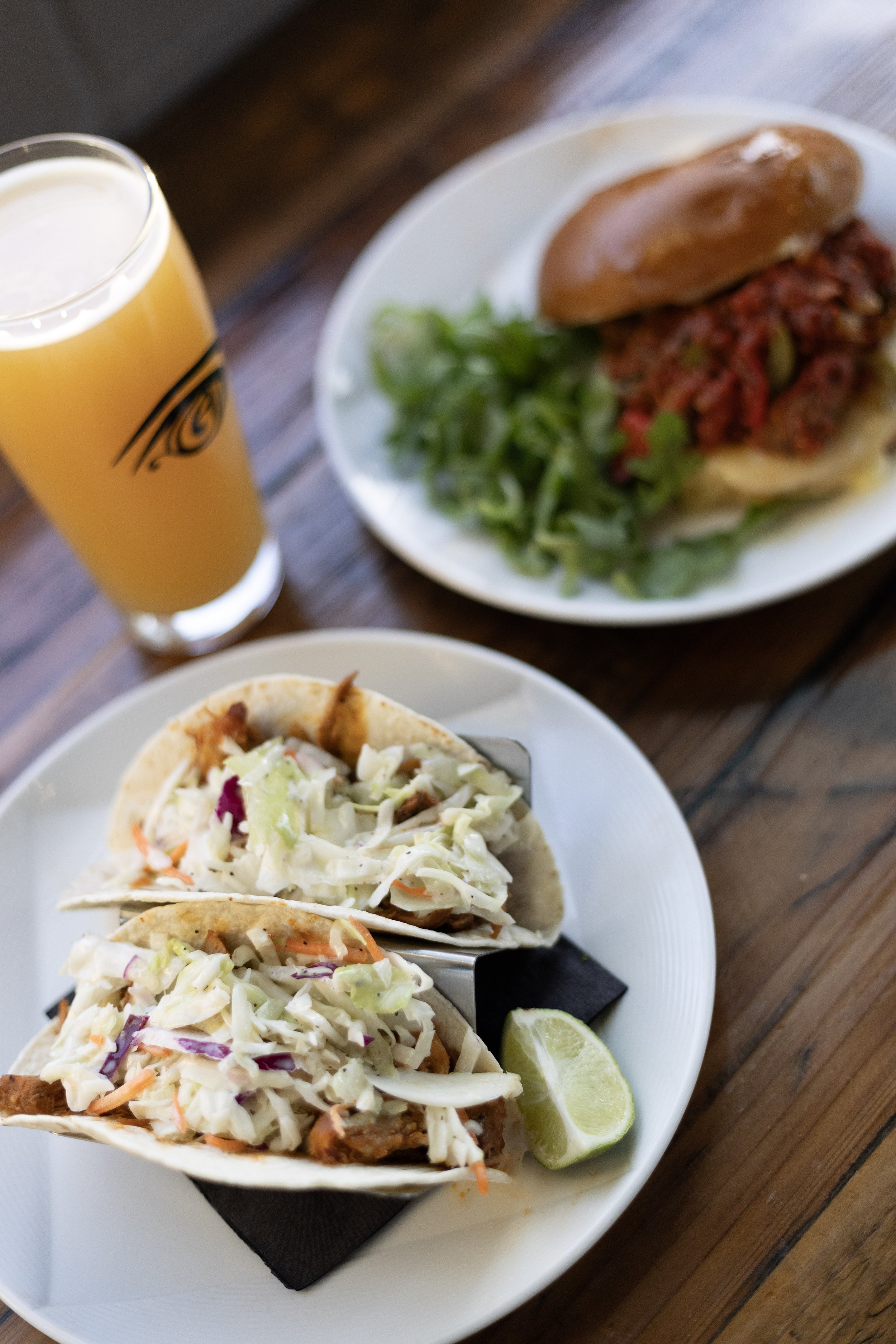 A plate with two tacos filled with shredded meat and topped with coleslaw, a wedge of lime, a plate with a sandwich containing meat, lettuce, and a bun, and a glass of orange juice on a wooden table.
