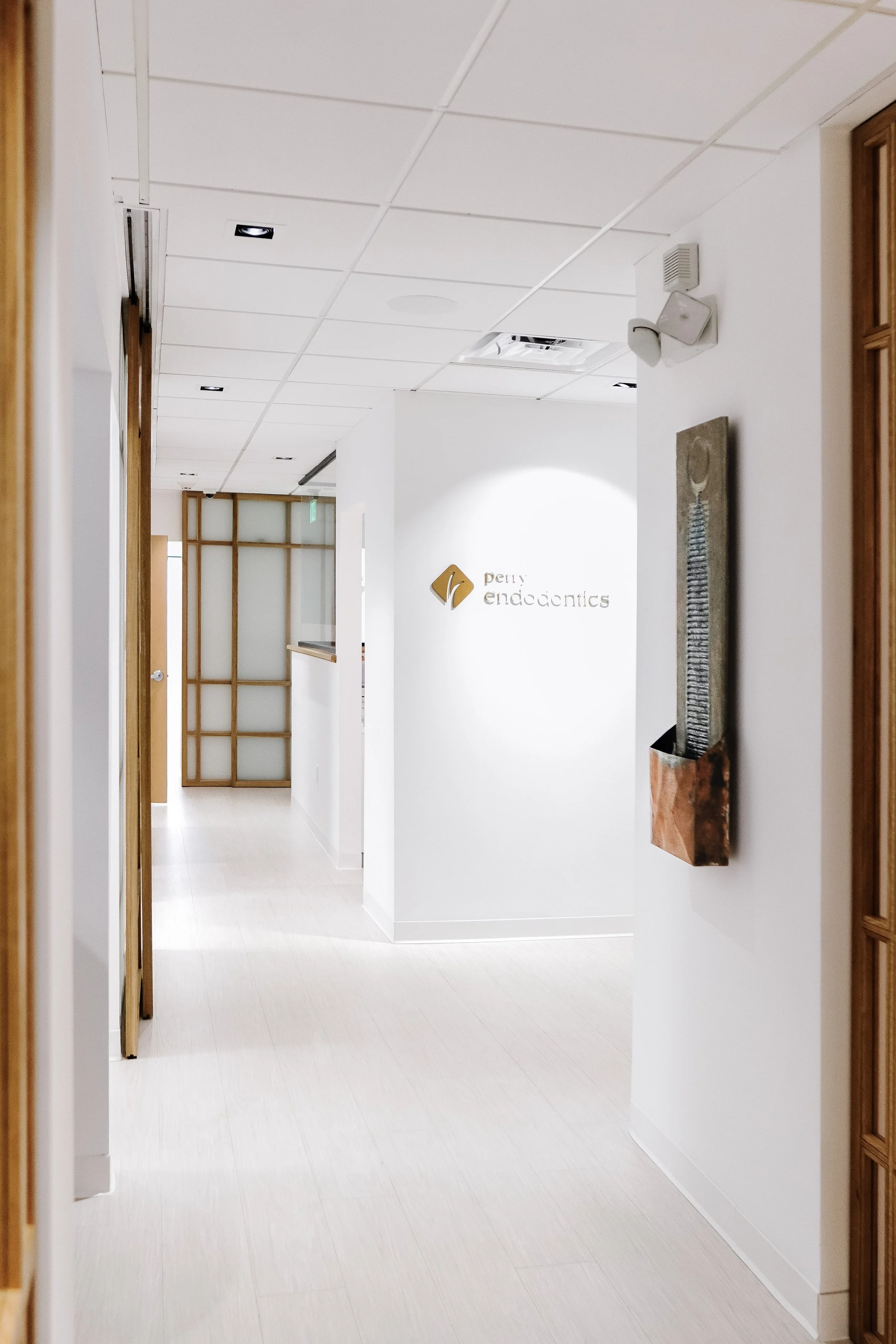 Interior view of a modern dental office hallway with white walls and light-colored wood accents, featuring a sign that reads 'Penny Endodontics' and abstract wall art.
