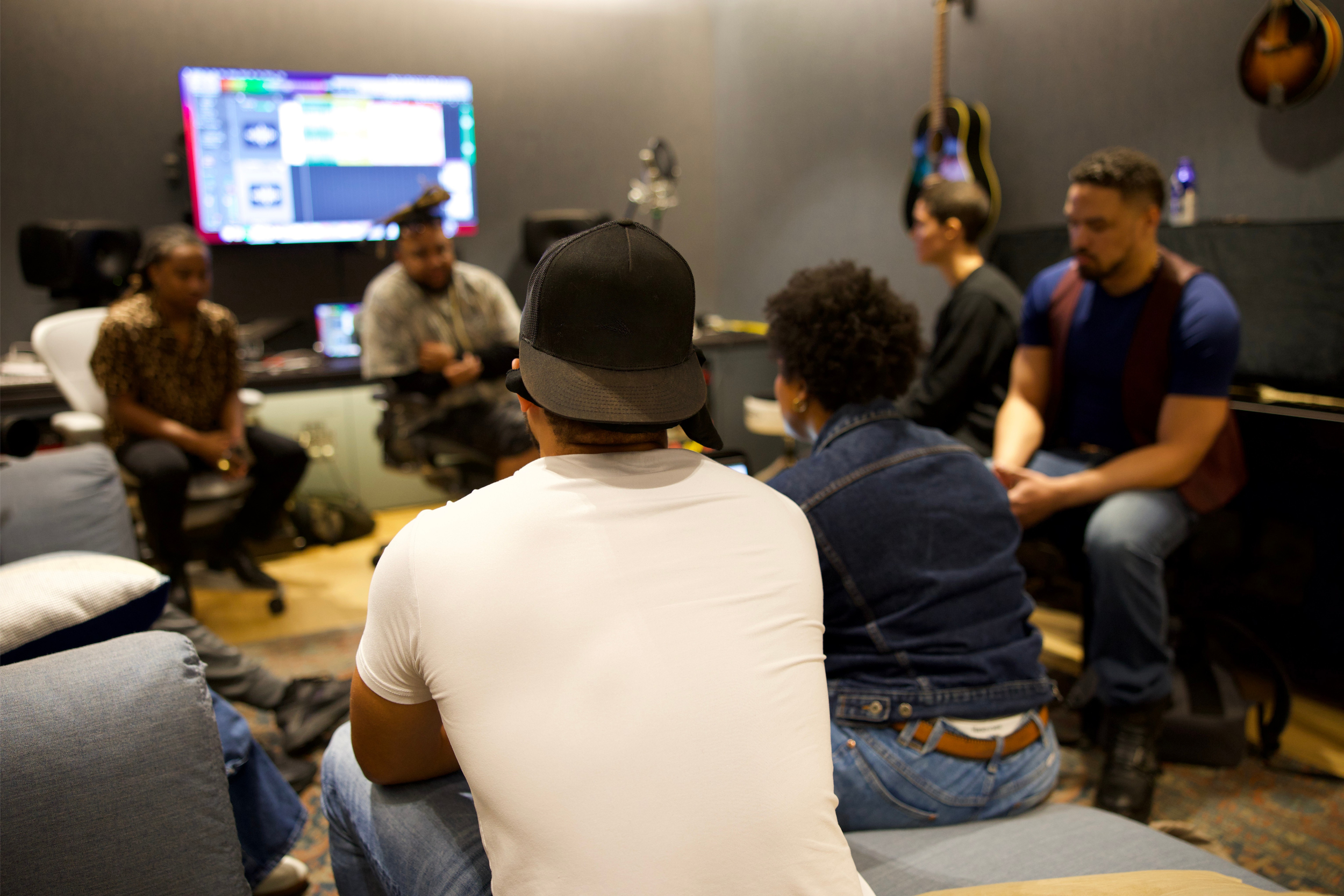 Group of people sitting and listening in a music studio with guitars hanging on the wall and a large screen display in the background.
