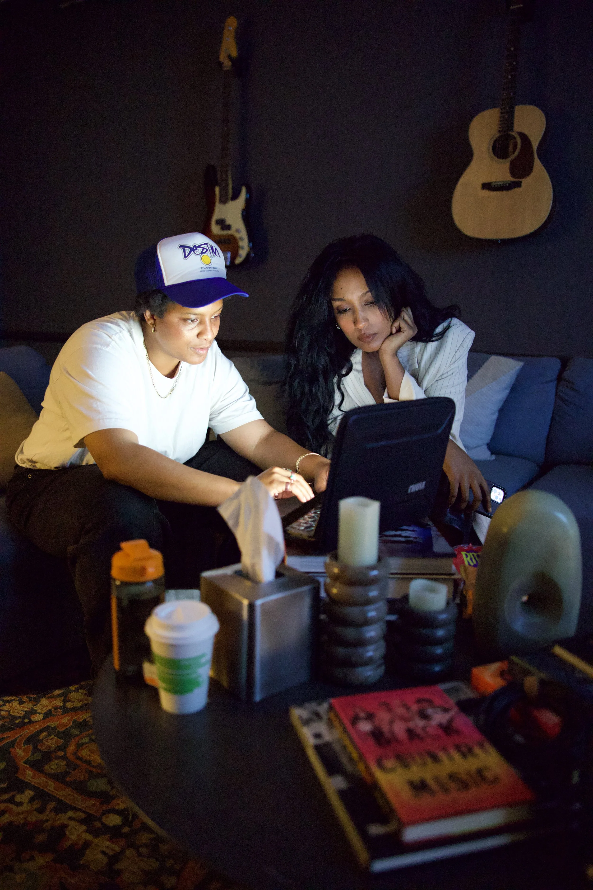Two women sitting on the floor in a dimly lit room, looking at a laptop screen. The woman on the left wears a white shirt and a baseball cap, while the woman on the right has long black hair and is wearing a striped blazer. A guitar and a bass guitar are hanging on the wall behind them, and there are various objects on a table in front of them.
