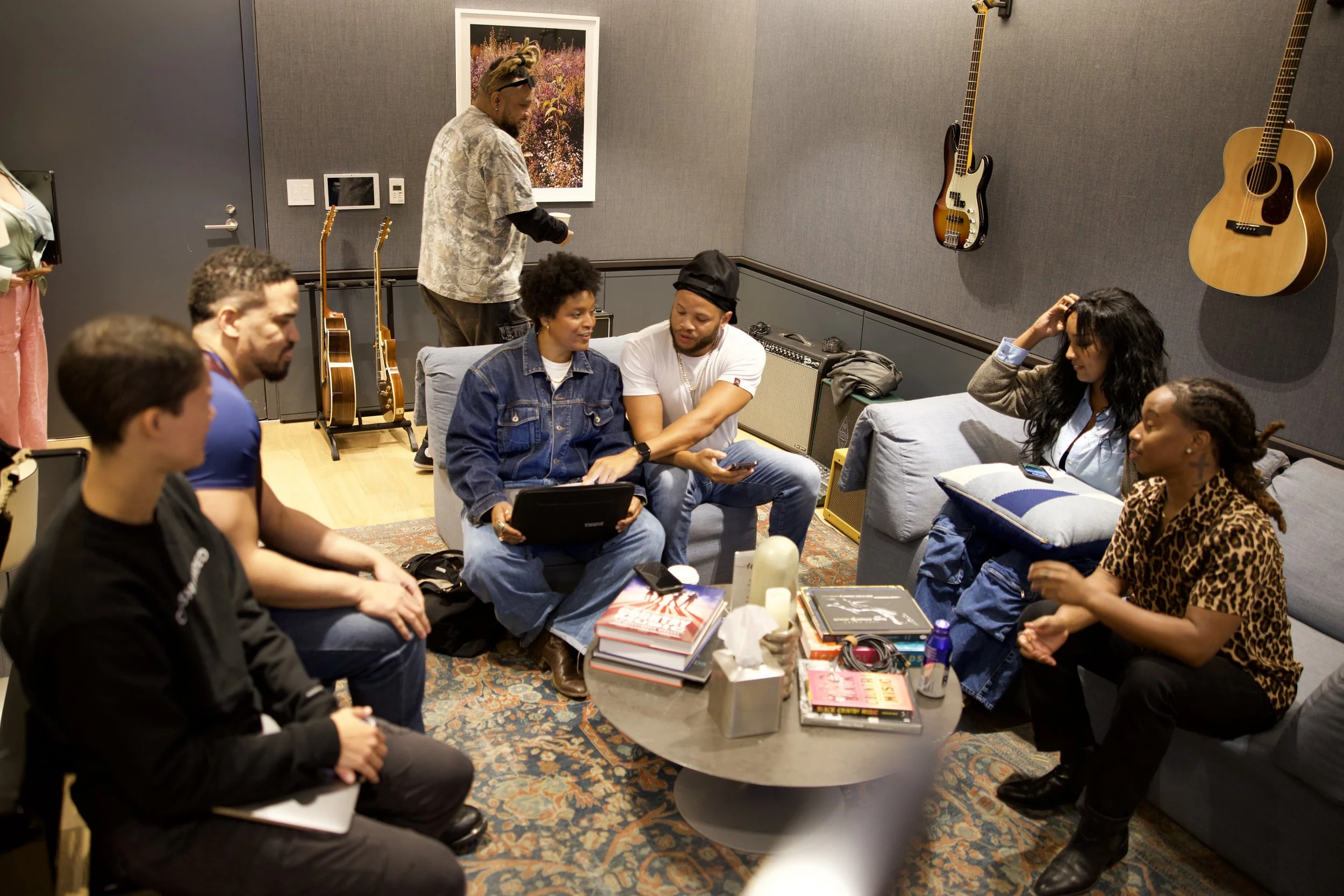 A group of people sitting around a coffee table with books, electronics, and drinks, some engaged in conversation, others looking at devices, in a room with guitars on the wall and framed artwork.