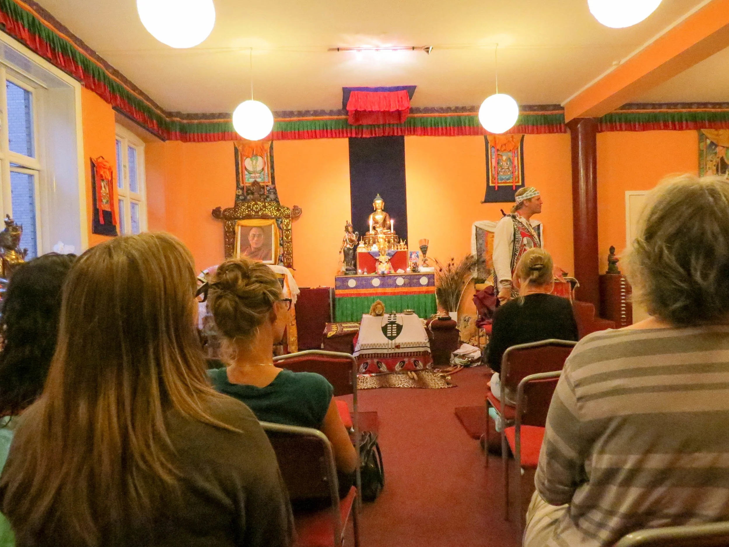 A group of people seated facing a spiritual or cultural presentation in a decorated room with orange walls, colorful banners, and religious artifacts on display.