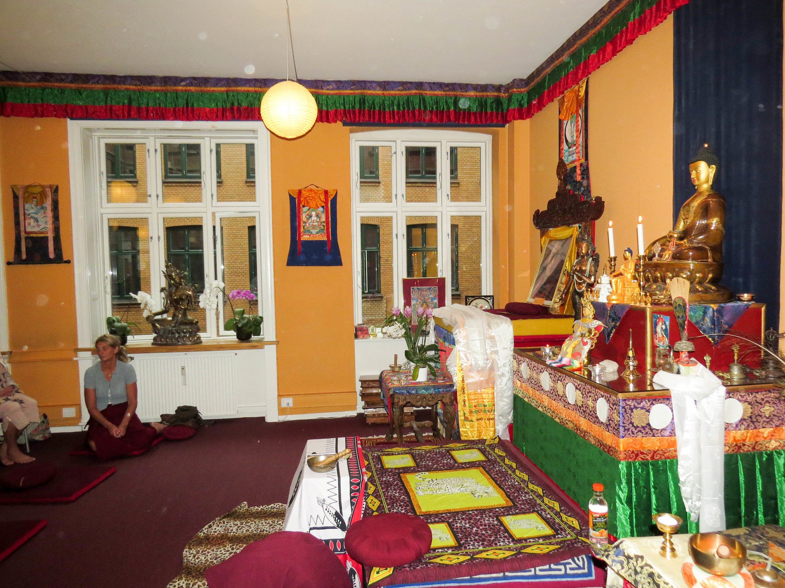 A room decorated with colorful Tibetan Buddhist altar statues, candles, and religious paintings. Two people sit on the floor in prayer or meditation.