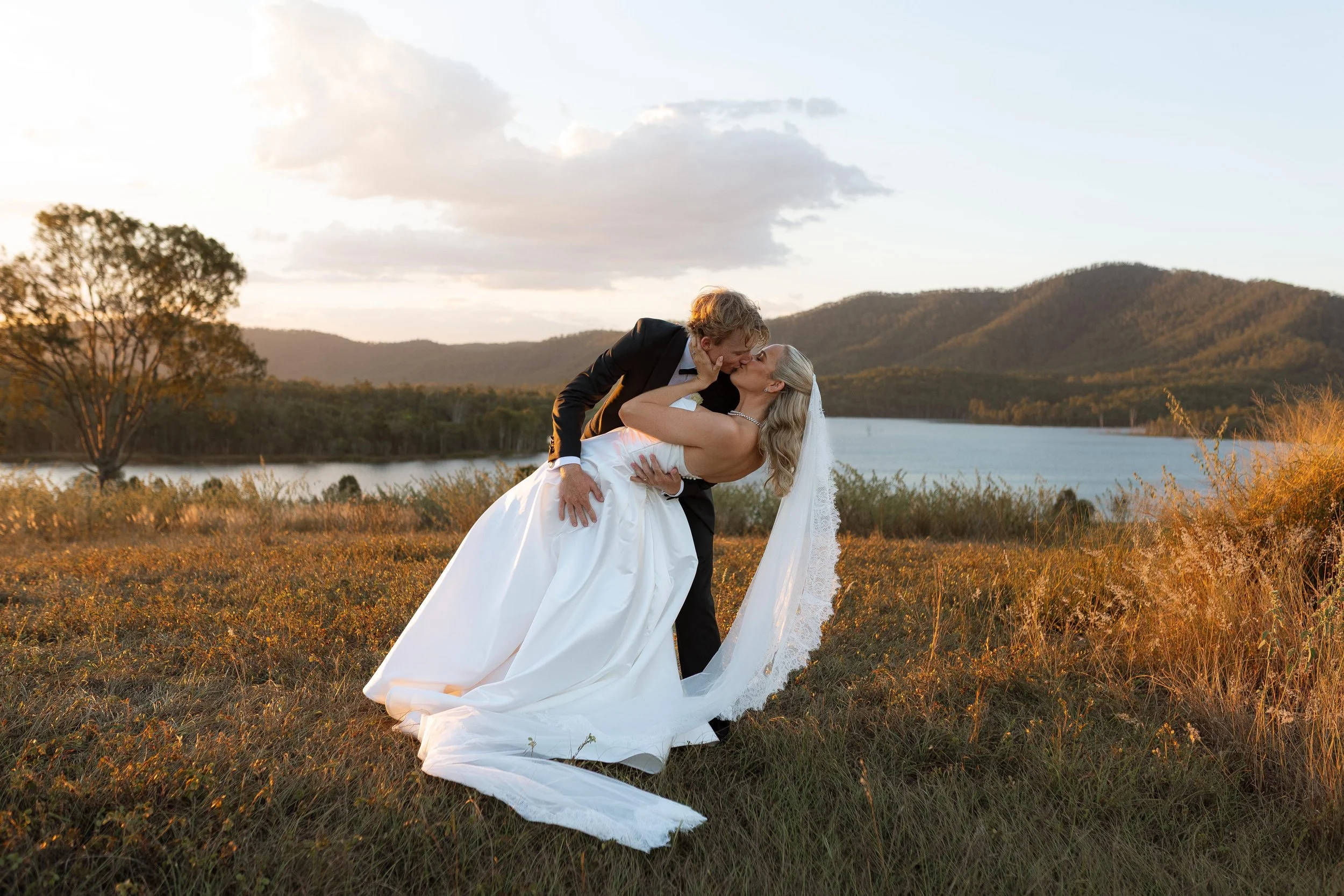 A bride and groom share a kiss outdoors with mountains, a lake, and trees in the background during sunset.