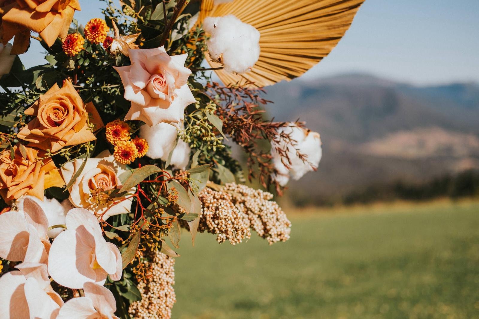 A floral arrangement featuring roses, marigolds, and other flowers with a large yellow paper parasol in the background, set against a scenic outdoor landscape with hills or mountains in the distance.