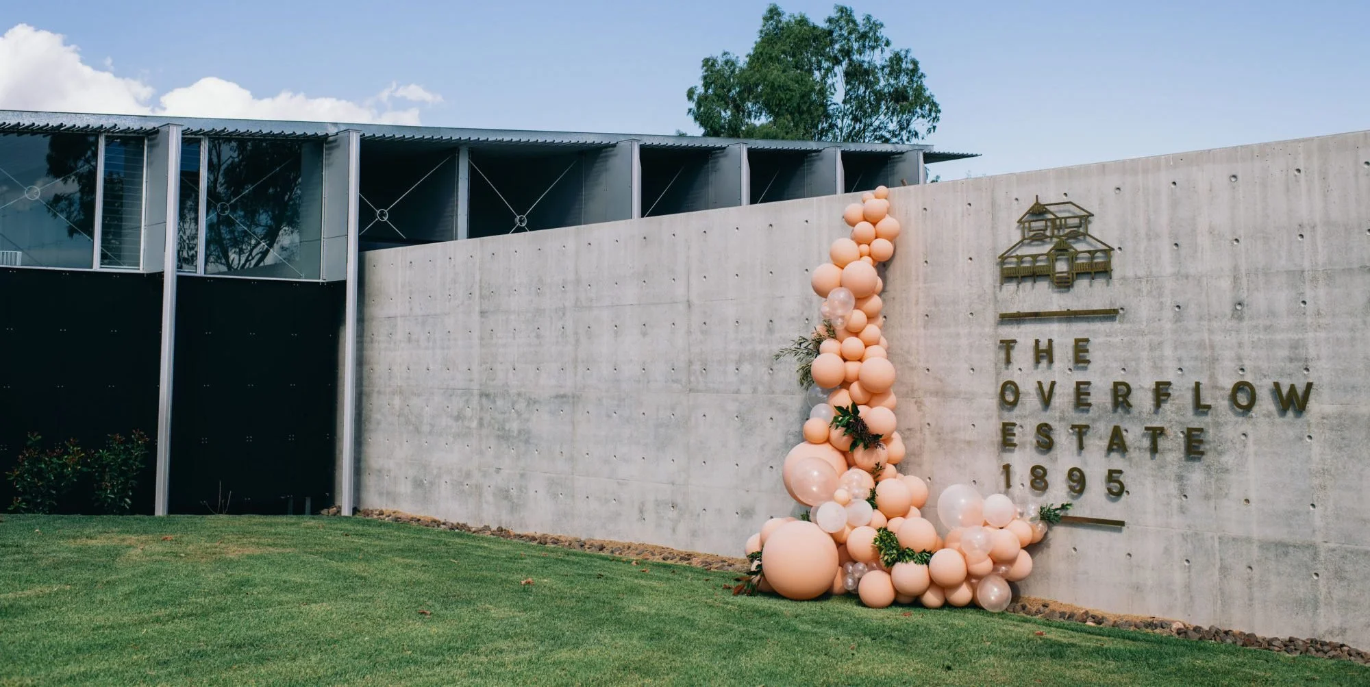 Modern building with metallic and glass exterior, decorated with pink balloons and greenery in front of a concrete wall that reads 'The Overflow Estate 1895' and features a small gold-colored house sculpture.