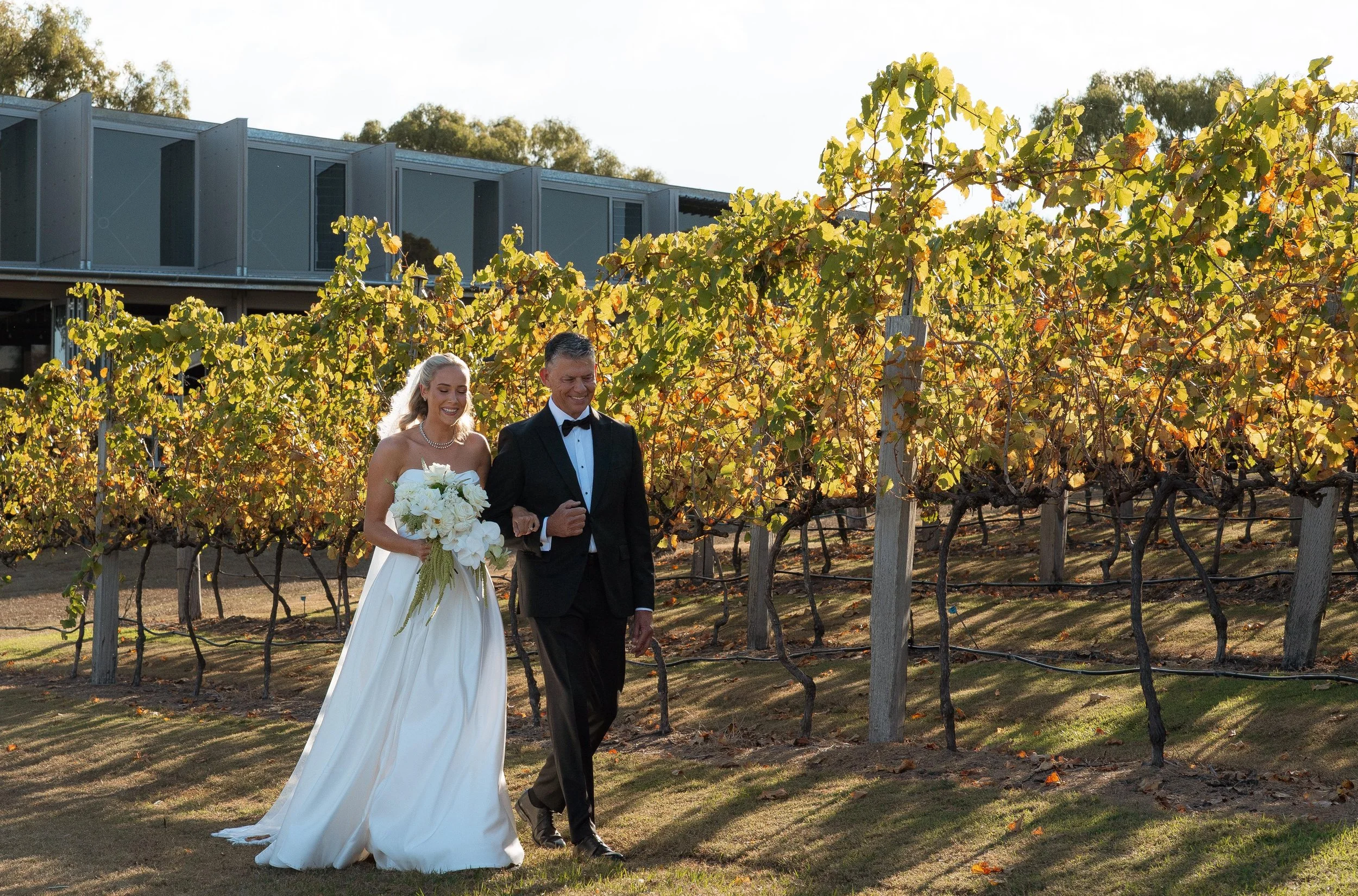 A bride and groom walk through a vineyard during a wedding celebration under bright sunlight, with trees and a modern building in the background.