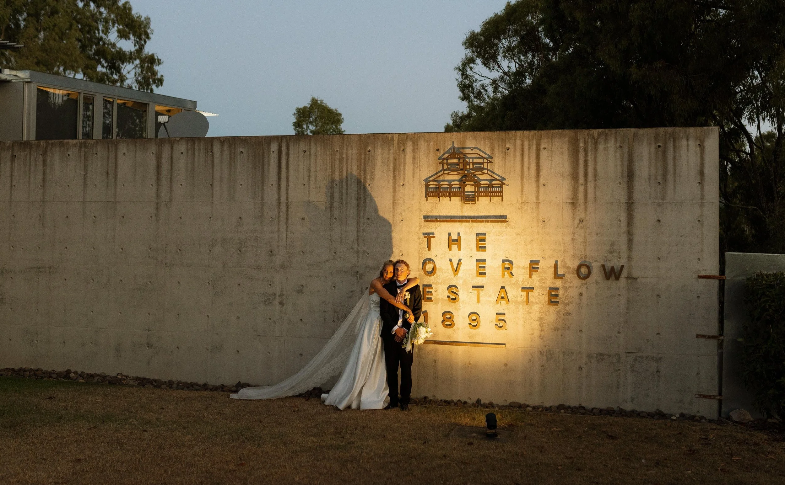 A couple dressed in wedding attire, standing in front of a large concrete wall with the text 'The Overflow Estate 1895' and a graphic of a house. The couple is embracing each other, with the bride wearing a white wedding gown and the groom in a black